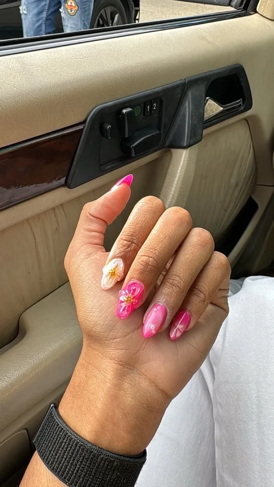Close-up of a hand with floral pink and white nail art, resting inside a car. The background shows part of the car door with window controls and beige interior upholstery.