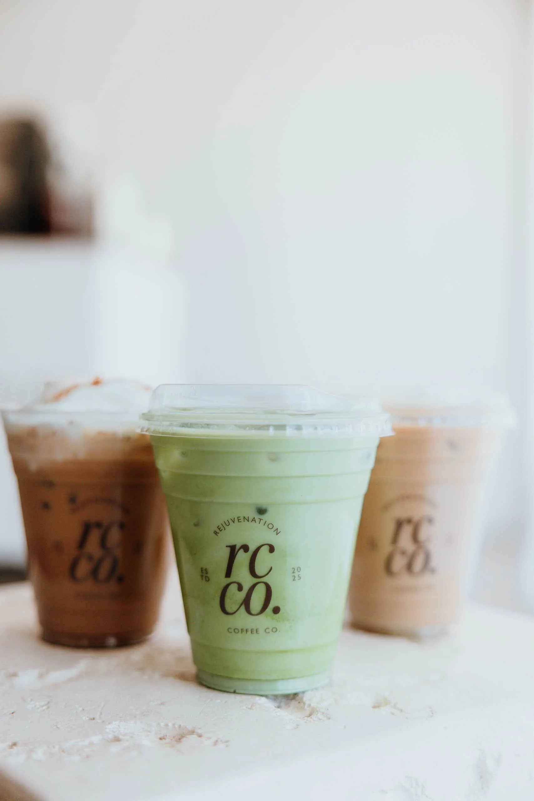 Three cups of iced coffee drinks on a white textured surface, with the focus on a green iced matcha latte in the front, flanked by two iced coffee beverages in brown cups, all with clear plastic lids.
