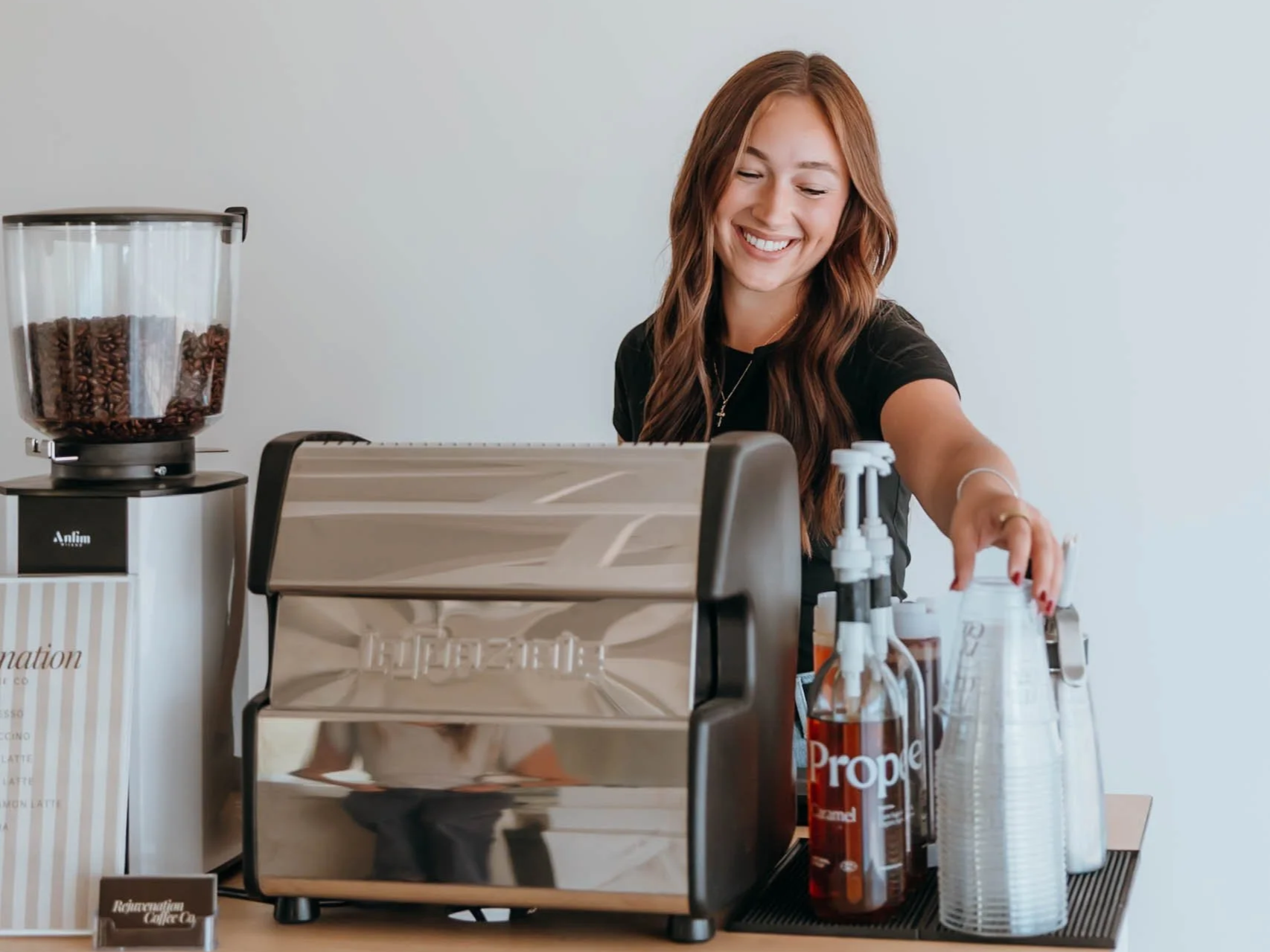 A smiling woman working behind a coffee or espresso machine with coffee supplies and cups on a table.