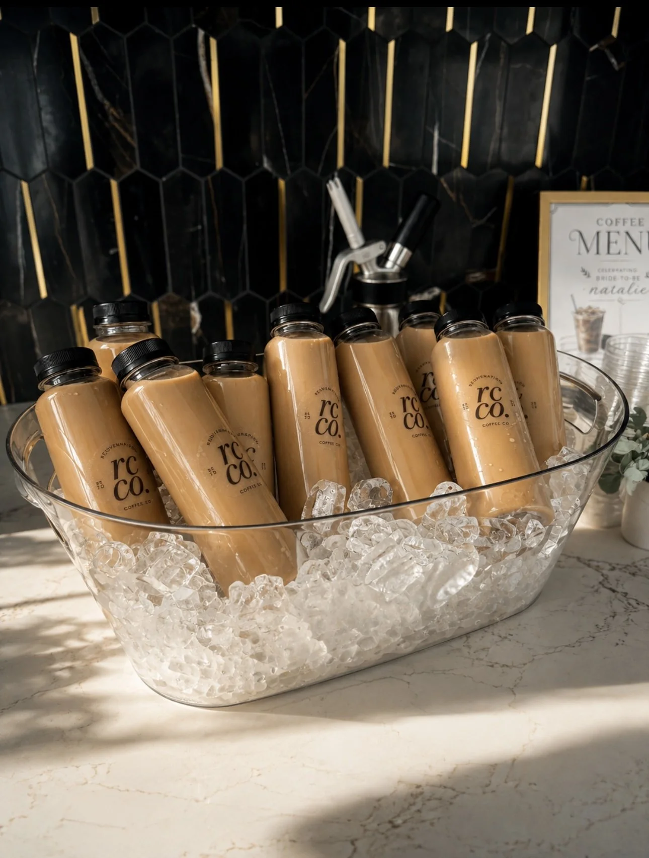 Glass bowl filled with ice and several bottled lattes. The background shows a black and gold hexagonal tile wall and a coffee station with dispensers.
