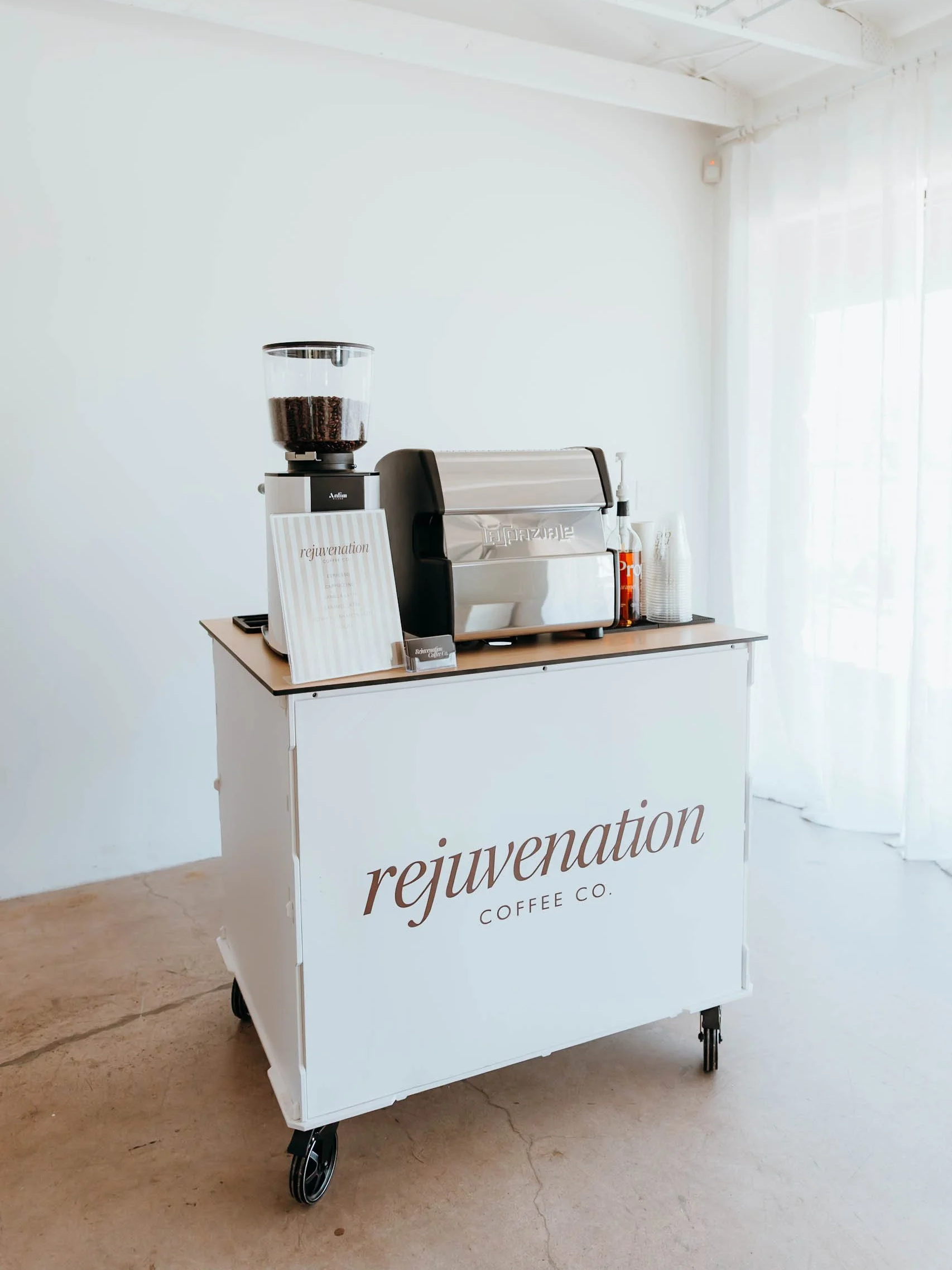 A white coffee cart on wheels labeled 'rejuvenation coffee co.' with a glass container of coffee beans, a black espresso machine, and disposable cups on top, set against a white wall with sheer curtains.