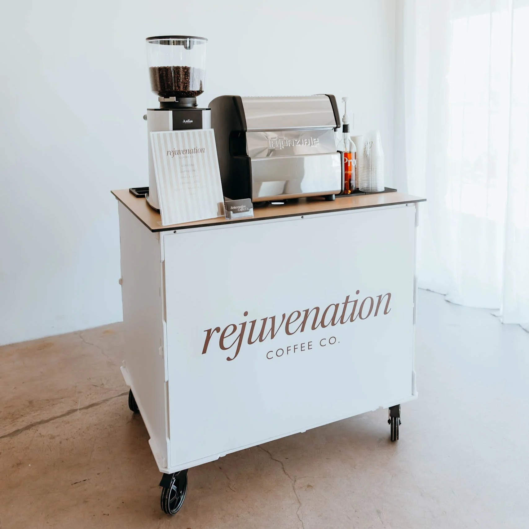 A white coffee cart on wheels with the name 'rejuvenation' and 'coffee co.' printed on the front. The cart has an espresso machine, a coffee grinder, and several paper cups.