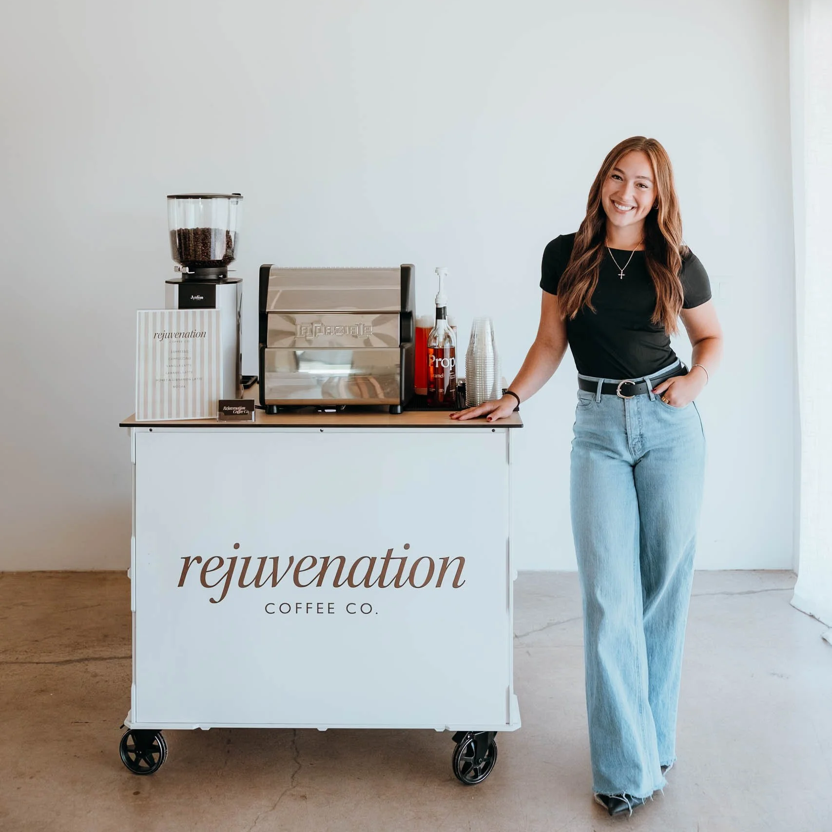 A professional barista standing next to a coffee stand with the words "rejuvenation coffee co." written on it, inside a minimalistic setting with white walls and a concrete floor.