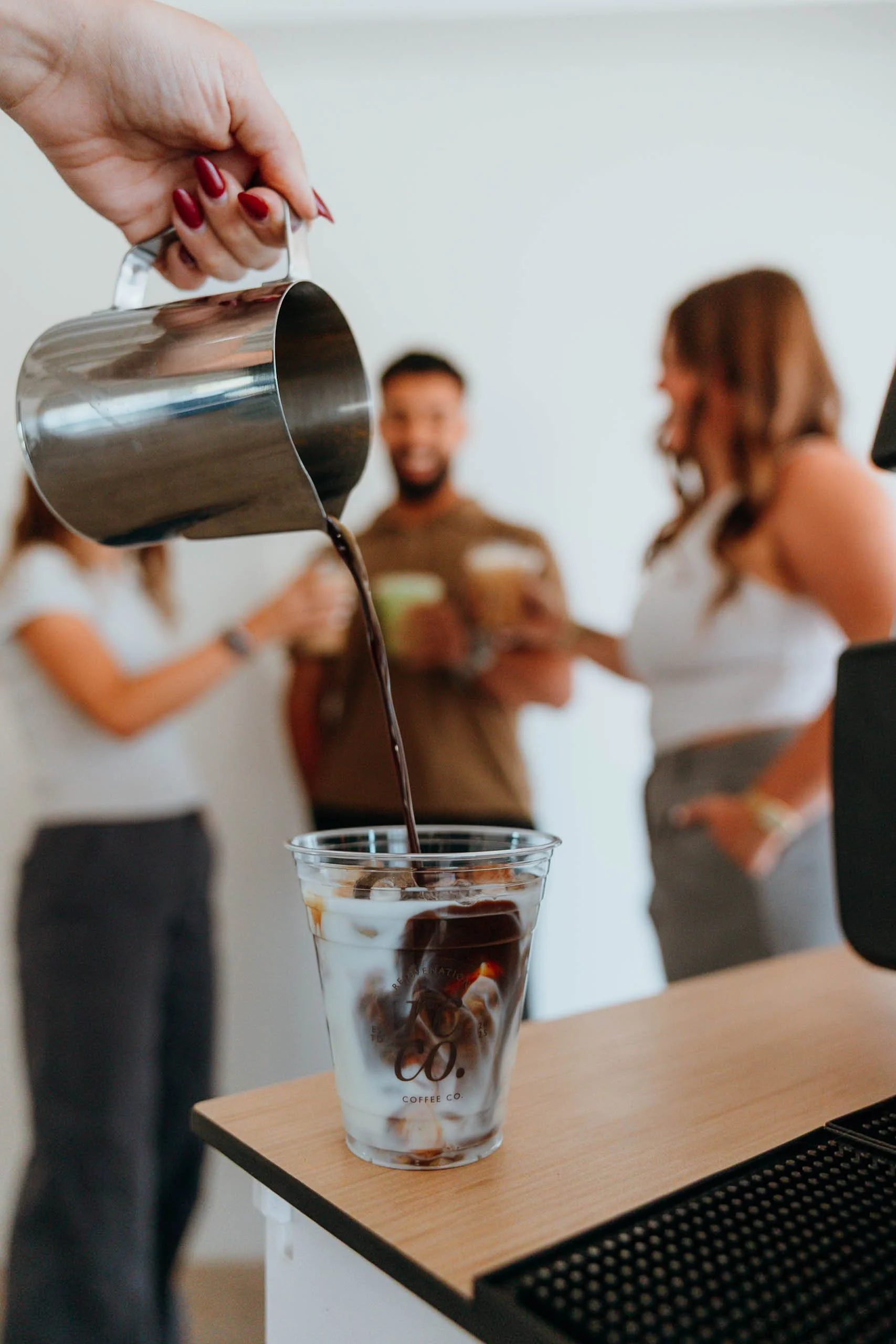 Pouring iced coffee into a clear glass at an event in Phoenix with people in the background