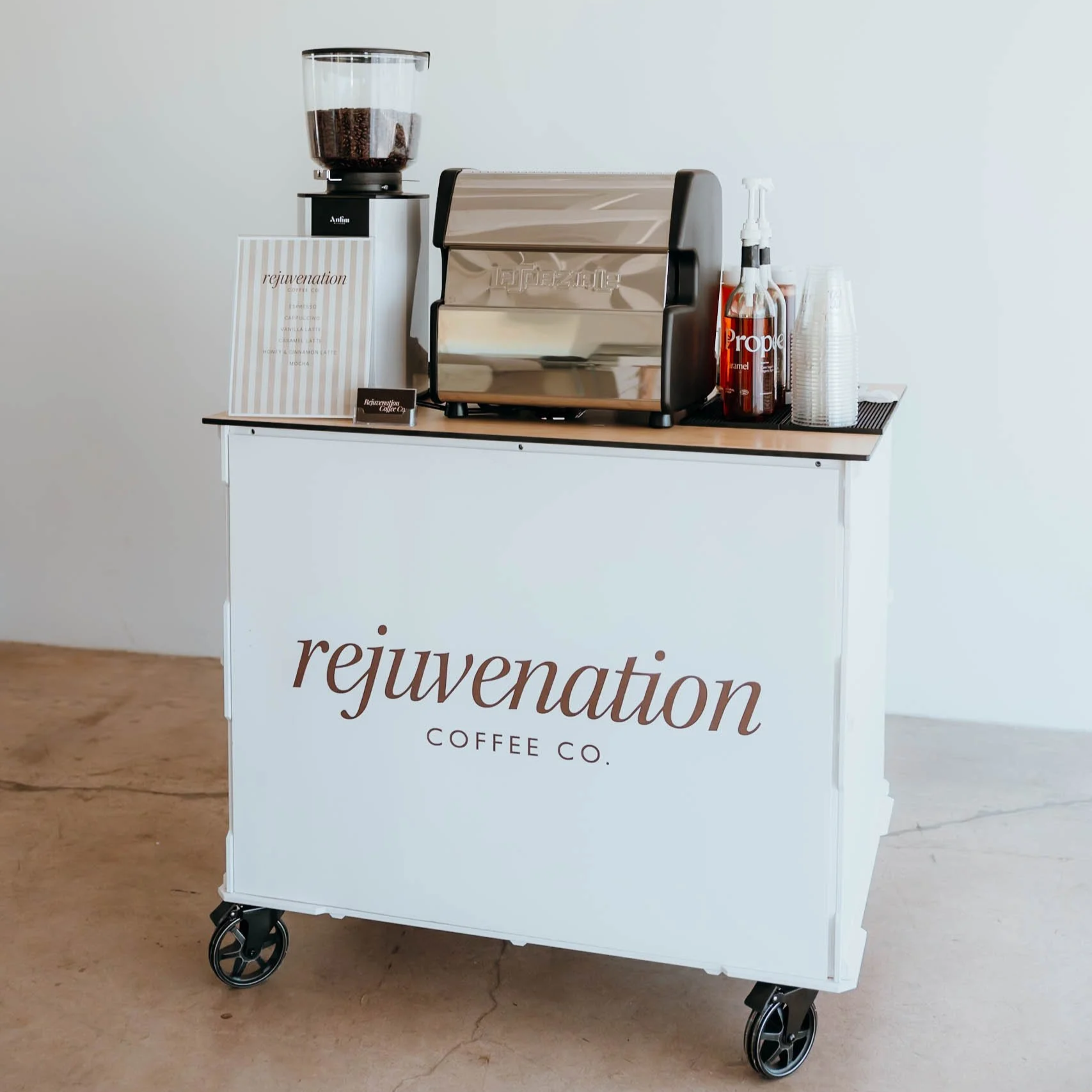 A mobile coffee cart with a white front panel displaying the text "rejuvenation COFFEE CO." A coffee grinder filled with coffee beans, a metallic espresso machine, and containers for syrup and cups are arranged on top of the cart. The background is plain white, and the floor is wood.