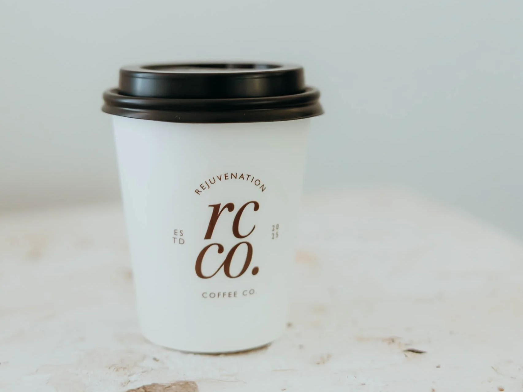 A white to-go coffee cup with a black plastic lid on a light-colored surface, featuring the logo of Rejuvenation Coffee Co.