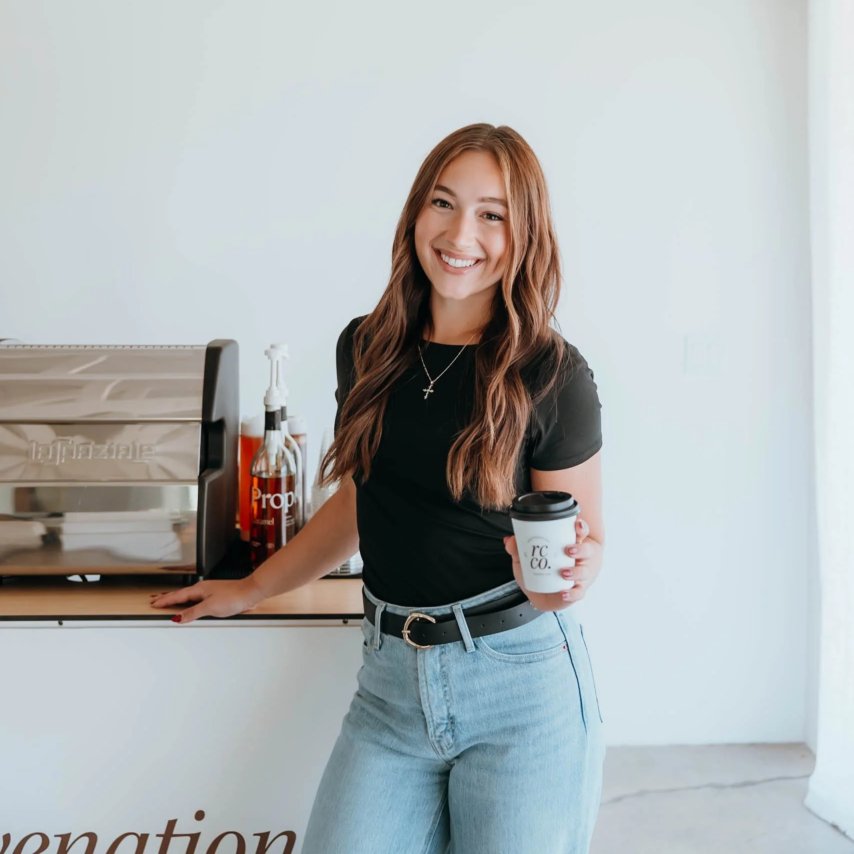 A woman with long, wavy, reddish-brown hair smiling and holding a coffee cup in a white cup holder. She is wearing a black top, light blue jeans, and a silver belt. She stands in front of a white wall with part of a coffee machine and some bottles in the background.
