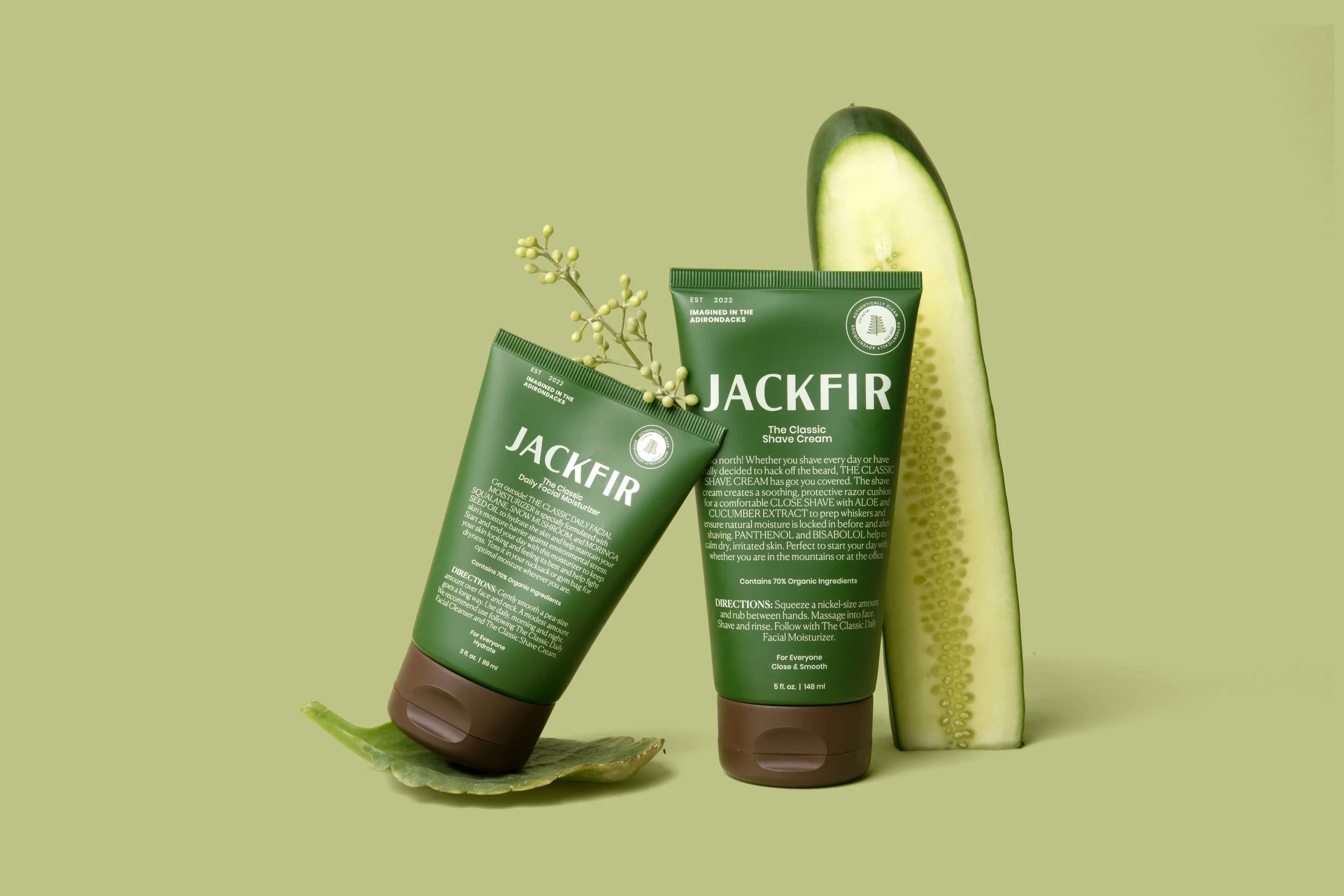 Two green tubes of Jackfir shave cream, a cucumber slice, and a small branch with buds on a green background.
