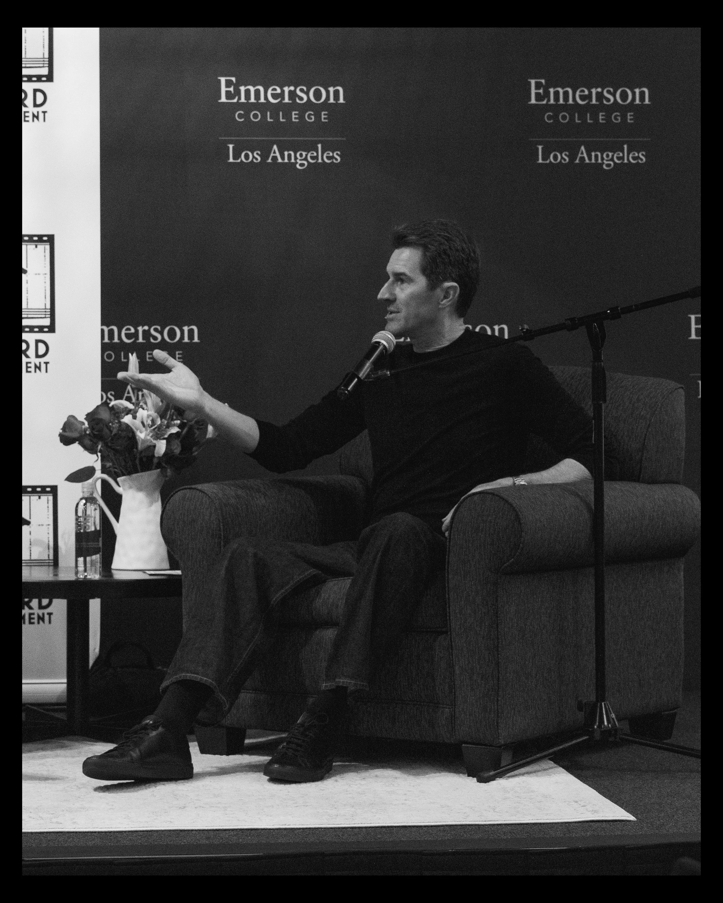 A man with dark hair and light skin sitting on a couch, wearing a black shirt and black shoes, speaking into a microphone in front of a black backdrop with the words "Emerson College Los Angeles" written on it. There is a table with a flower bouquet 