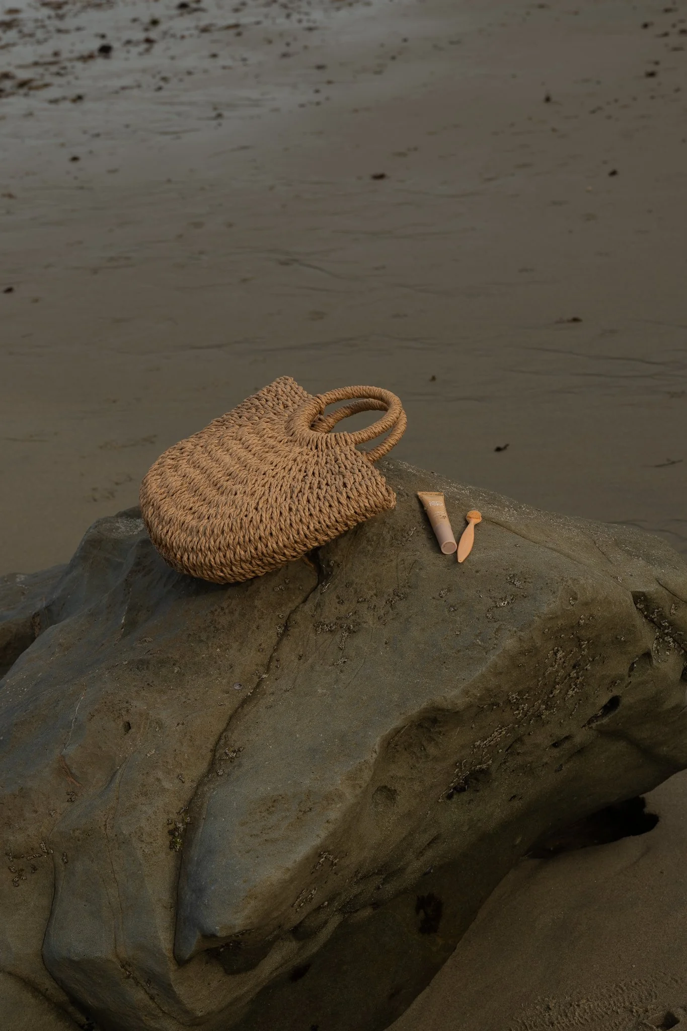 A woven straw handbag and skincare or makeup products on a rock near the beach.