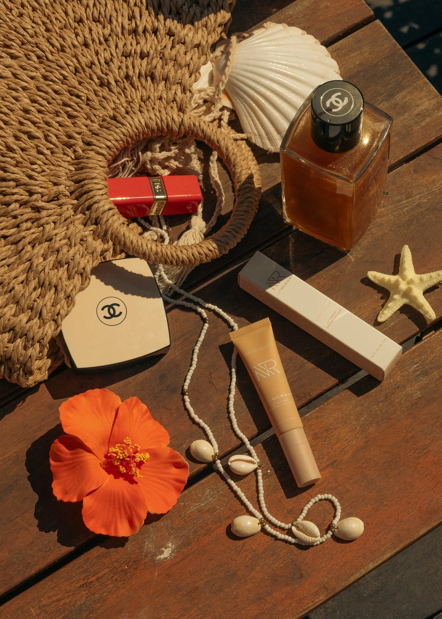 A collection of summer-themed items on a wooden surface, including a straw bag, a seashell, a bottle of perfume, a red lipstick, a starfish, a white box of beauty products, a twist of orange flower, and white jewelry with shells.