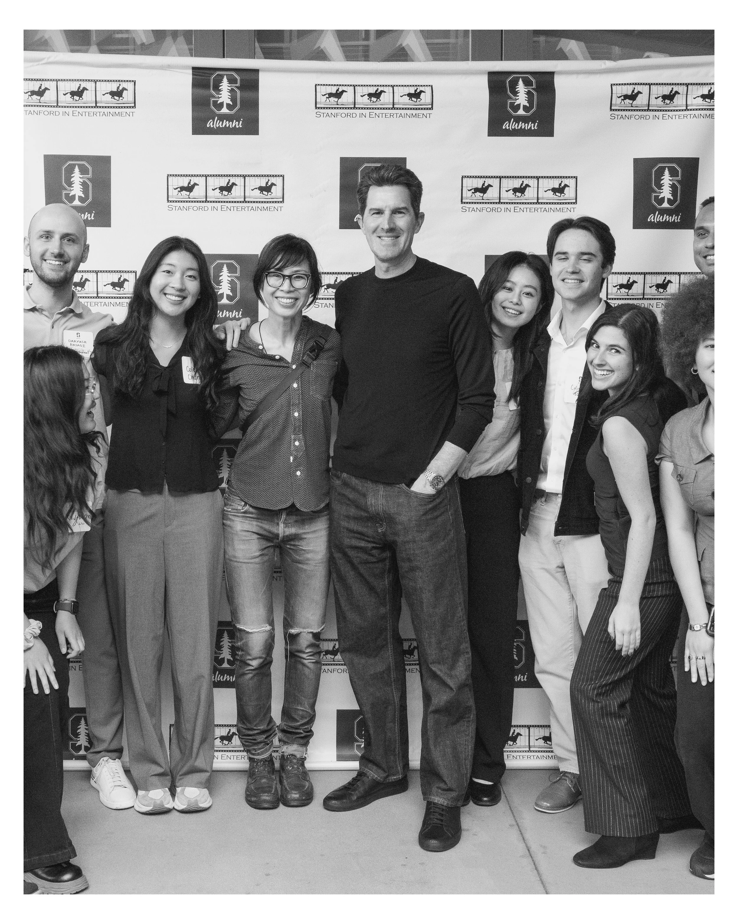 Group of people smiling and standing together at an event, with a backdrop displaying Stanford University logos and text, in black-and-white.