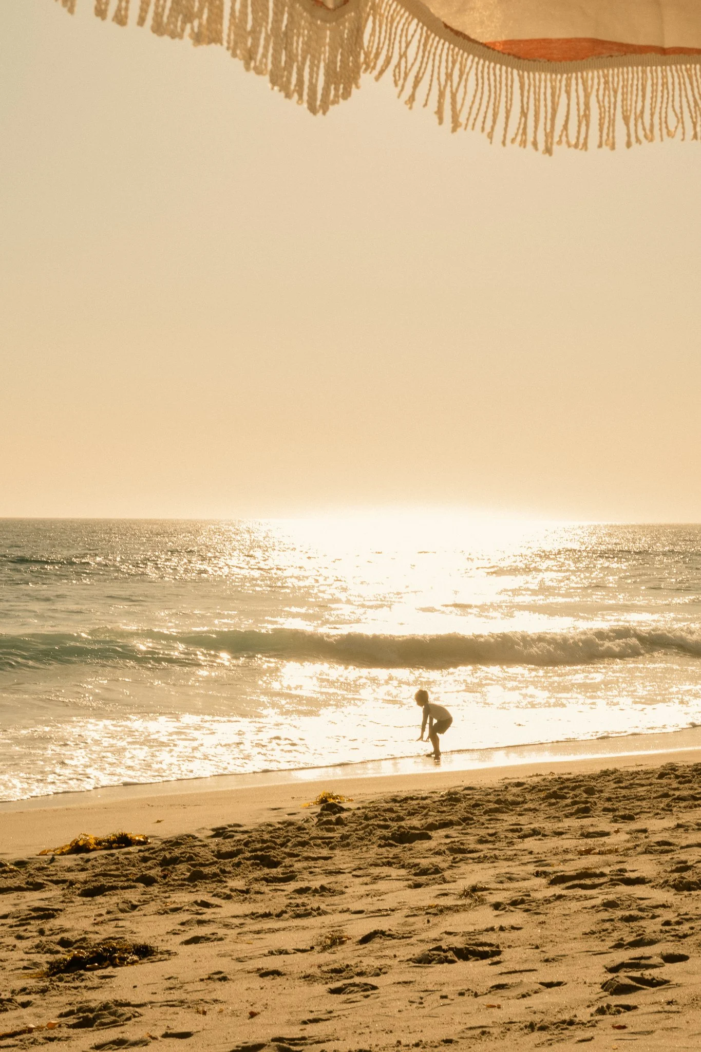 A person bending down on a sandy beach, with ocean waves in the background, and a fringed sail or curtain hanging overhead.