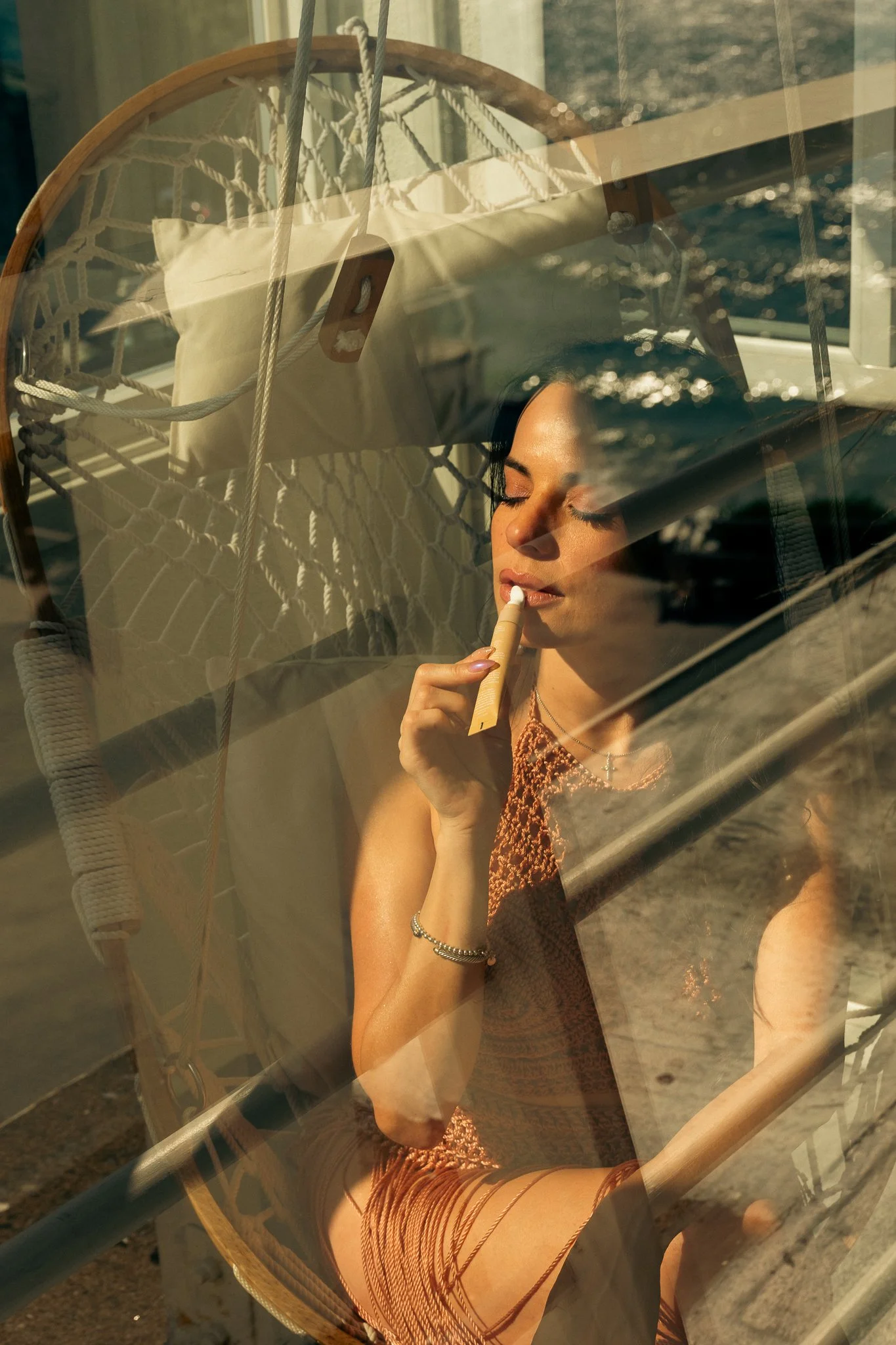A woman with dark hair and light skin is sitting and applying lipstick while sitting in front of a glass window, with an outdoor scene reflecting in the glass.