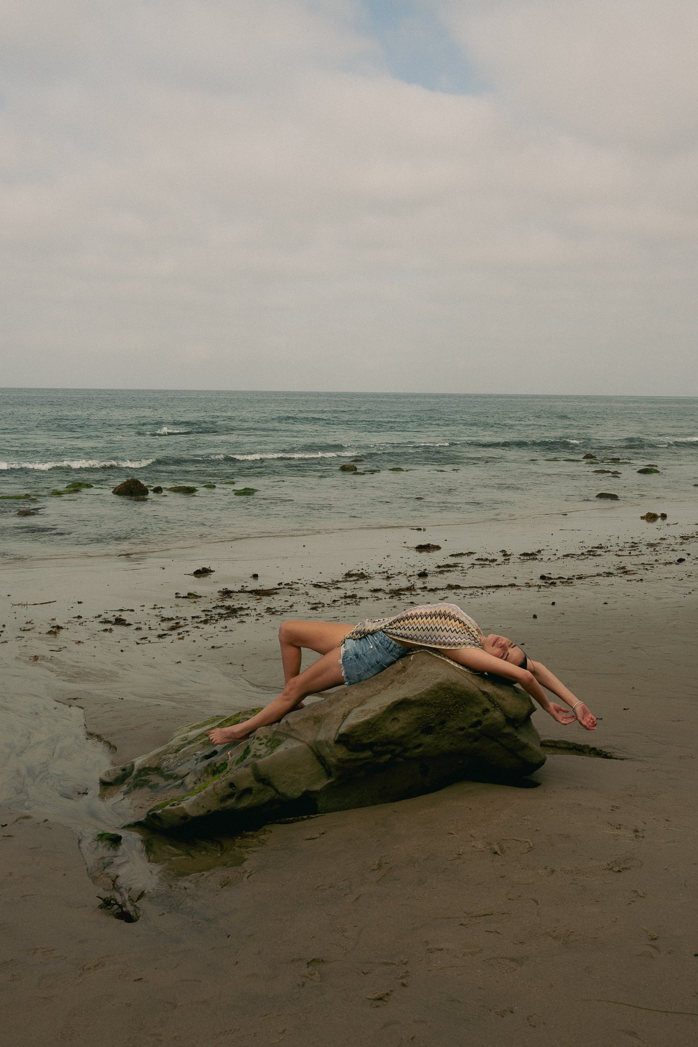 A young girl is lying on a large rock on a sandy beach, with her arm extended and her eyes closed. She is wearing a sleeveless top and denim shorts, and there are rocks scattered along the shoreline. The ocean and cloudy sky are in the background.