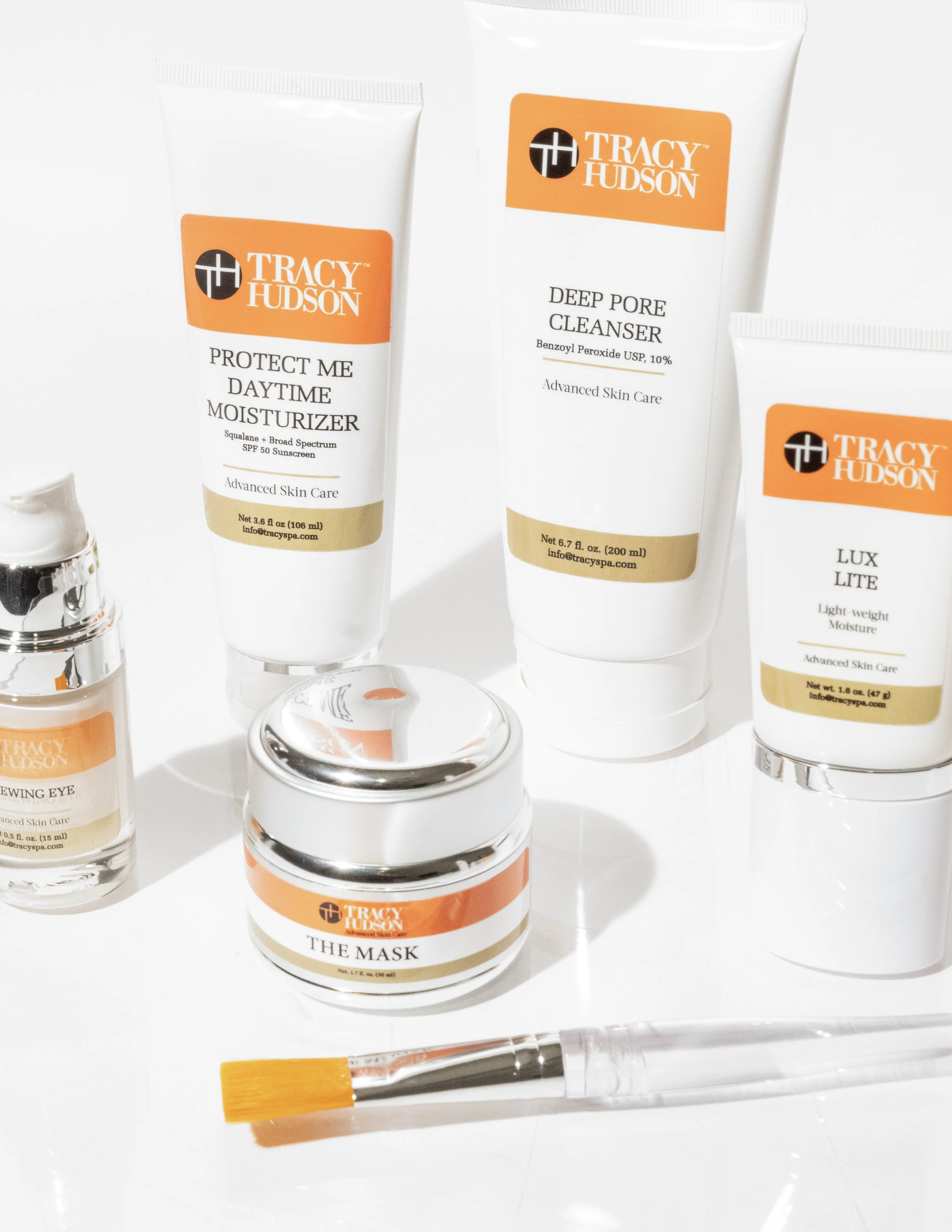 Collection of skincare and beauty products from Tracy Hudson, including a deep pore cleanser, a moisturizer named Protect Me Daytime Moisturizer, a mask, and a small jar, arranged on a white surface with a makeup brush below.