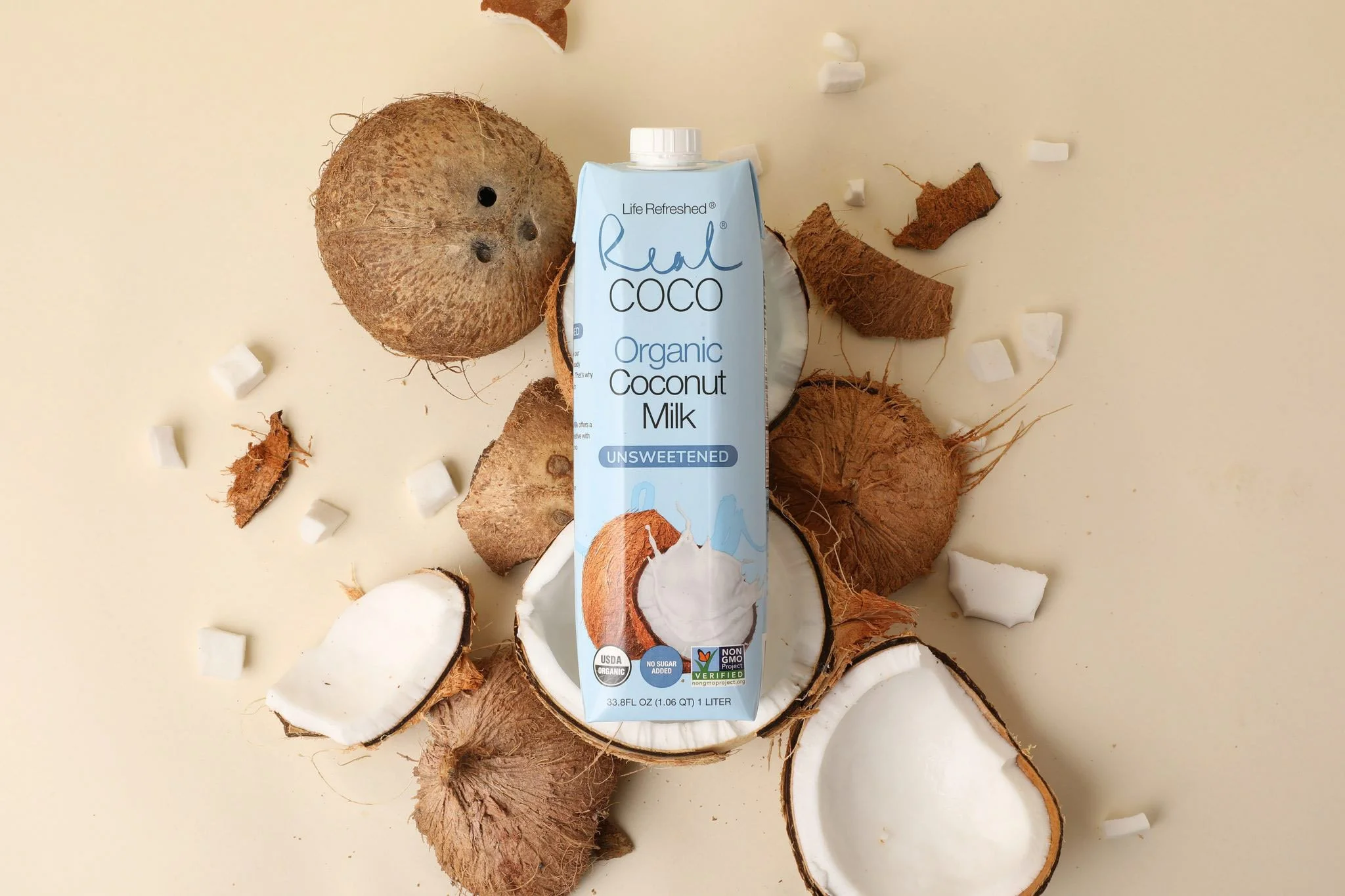 A carton of organic coconut milk standing upright on a light surface surrounded by halved and whole coconuts and coconut pieces.