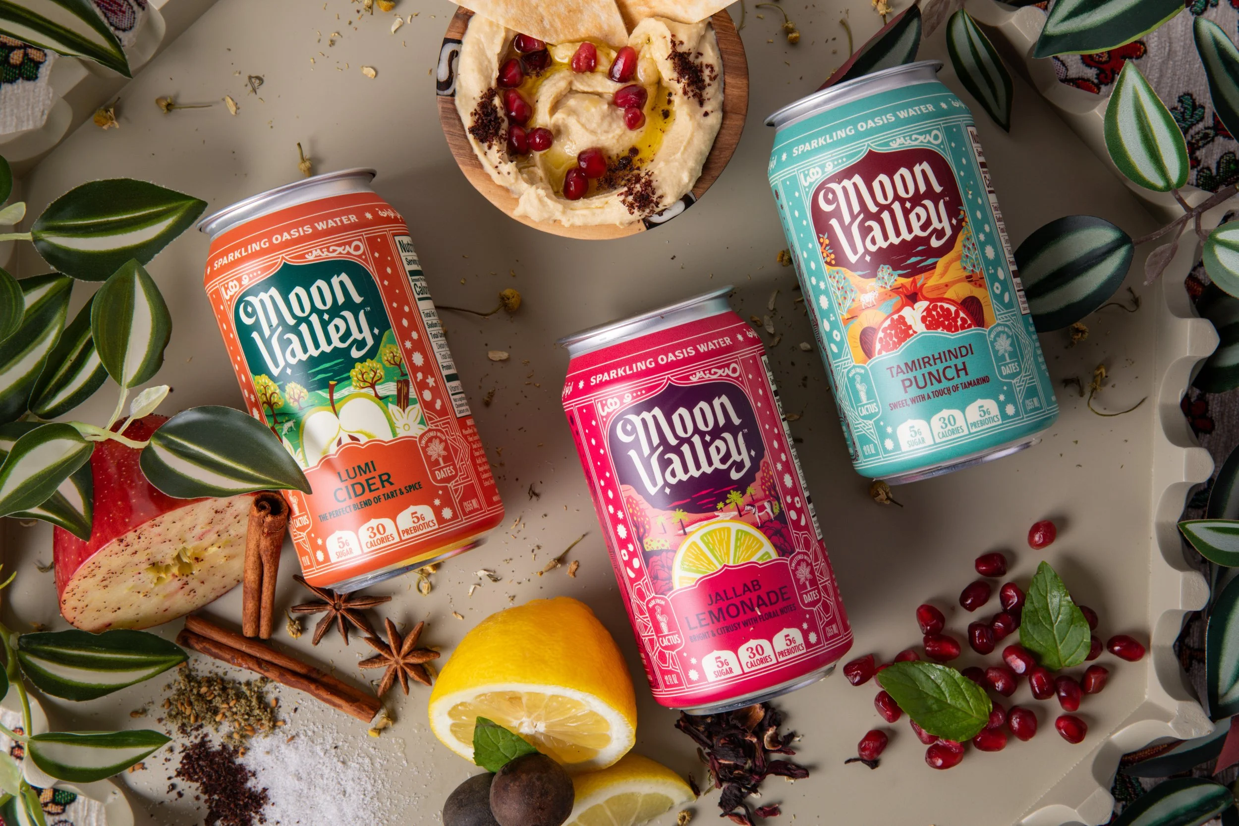 Arrangement of colorful Moon Valley drink cans with a bowl of hummus topped with pomegranate seeds and olive oil; lemon slices, apple, star anise, cinnamon sticks, and green leaves around the cans on a white surface.