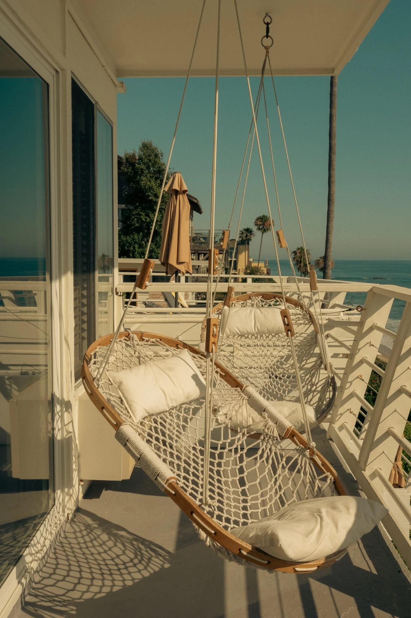 Balcony with wooden swing chair with cream cushions, shadow of the swing cast on the floor, glass railing, a beige umbrella, trees, buildings, and palm trees in the background with the ocean and sky visible.