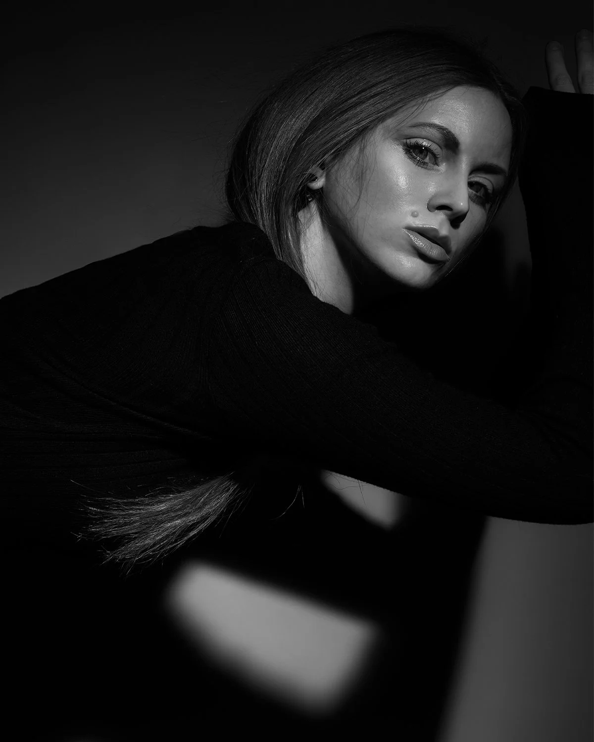 Black and white portrait of a woman with long hair, looking at the camera, with one arm raised, in low light.