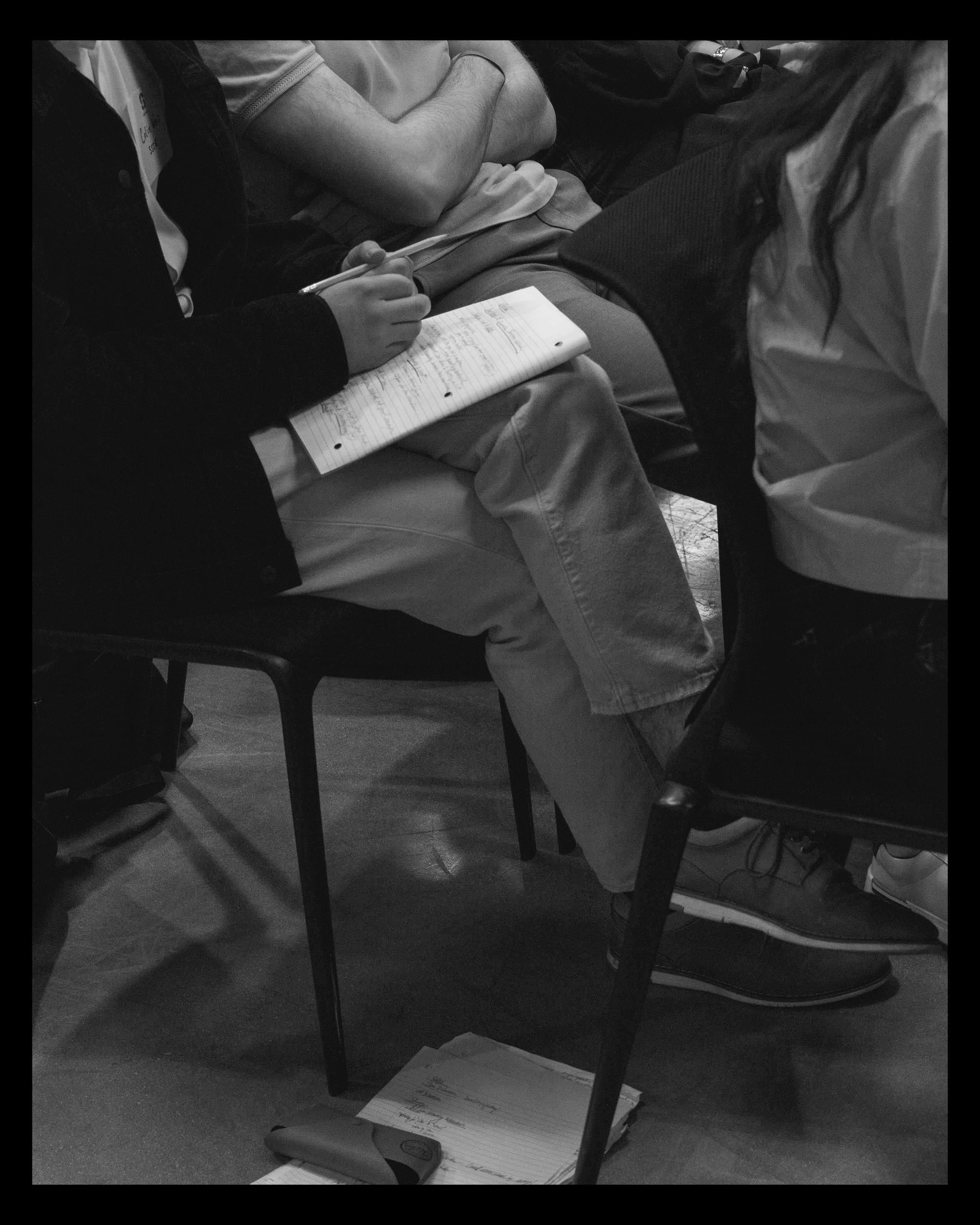 A person sitting on a chair with crossed arms wearing a short-sleeved shirt. They are taking notes in a spiral notebook with their right hand, with a pen in hand. A skateboard rests on the floor beneath their legs. Another person sits nearby, partial