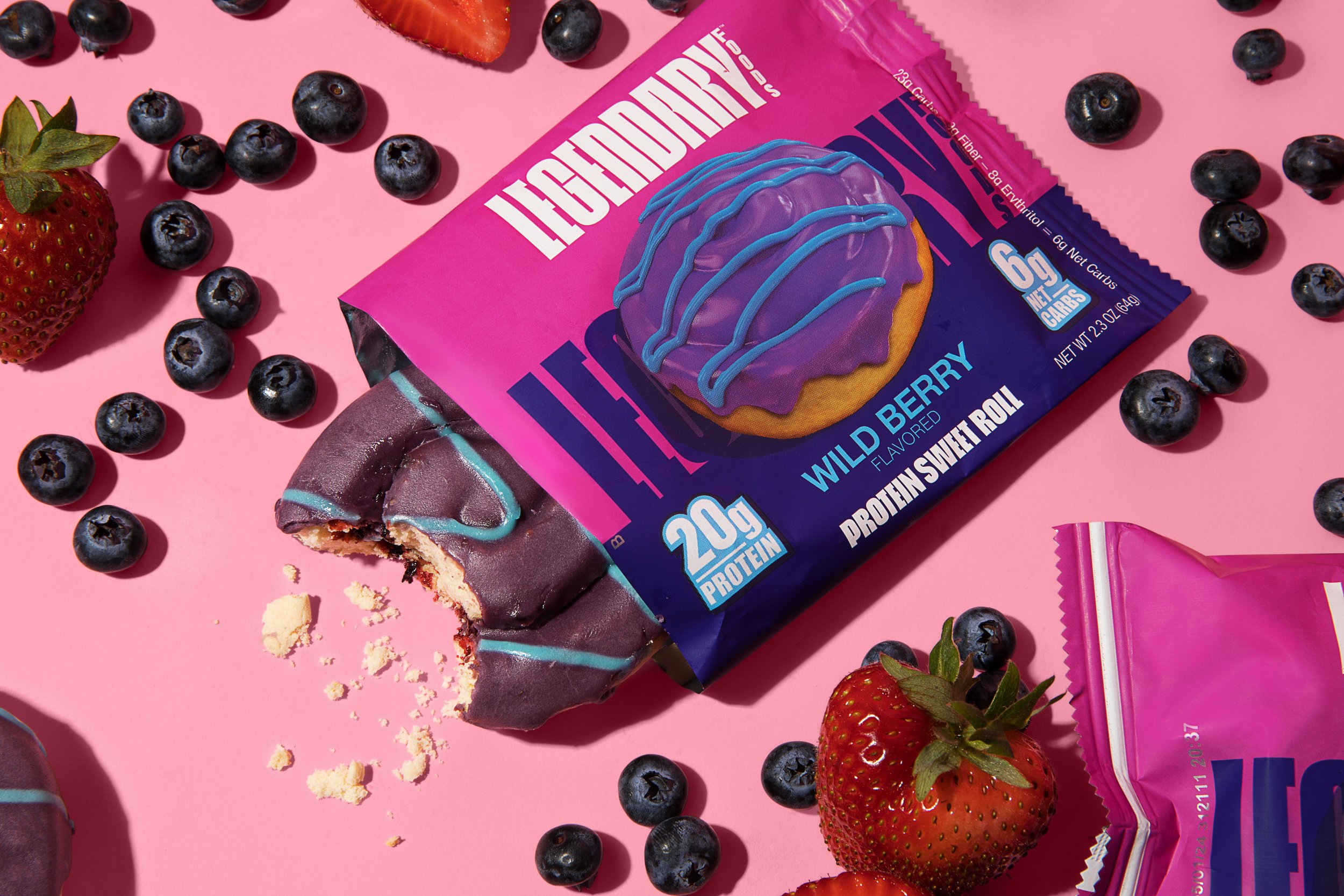 ProteinBuilder Prothen Sweet Roll protein snack bar in purple and pink packaging on pink background, surrounded by strawberries, blueberries, and crumbs.