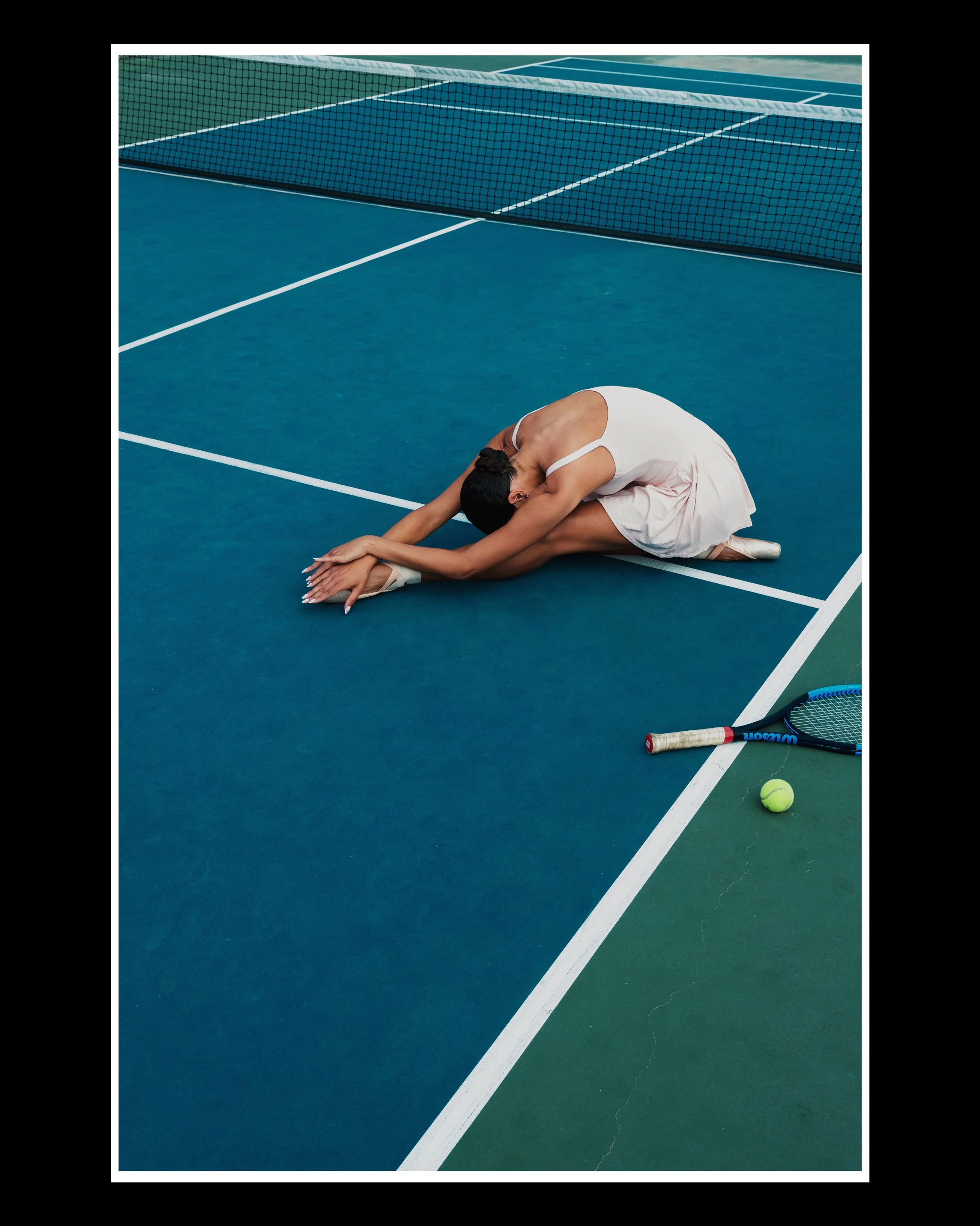 A female tennis player wearing a white dress and sitting on a tennis court with her head resting on her knees and arms stretched out in front. Several tennis rackets and a tennis ball are on the court near her.