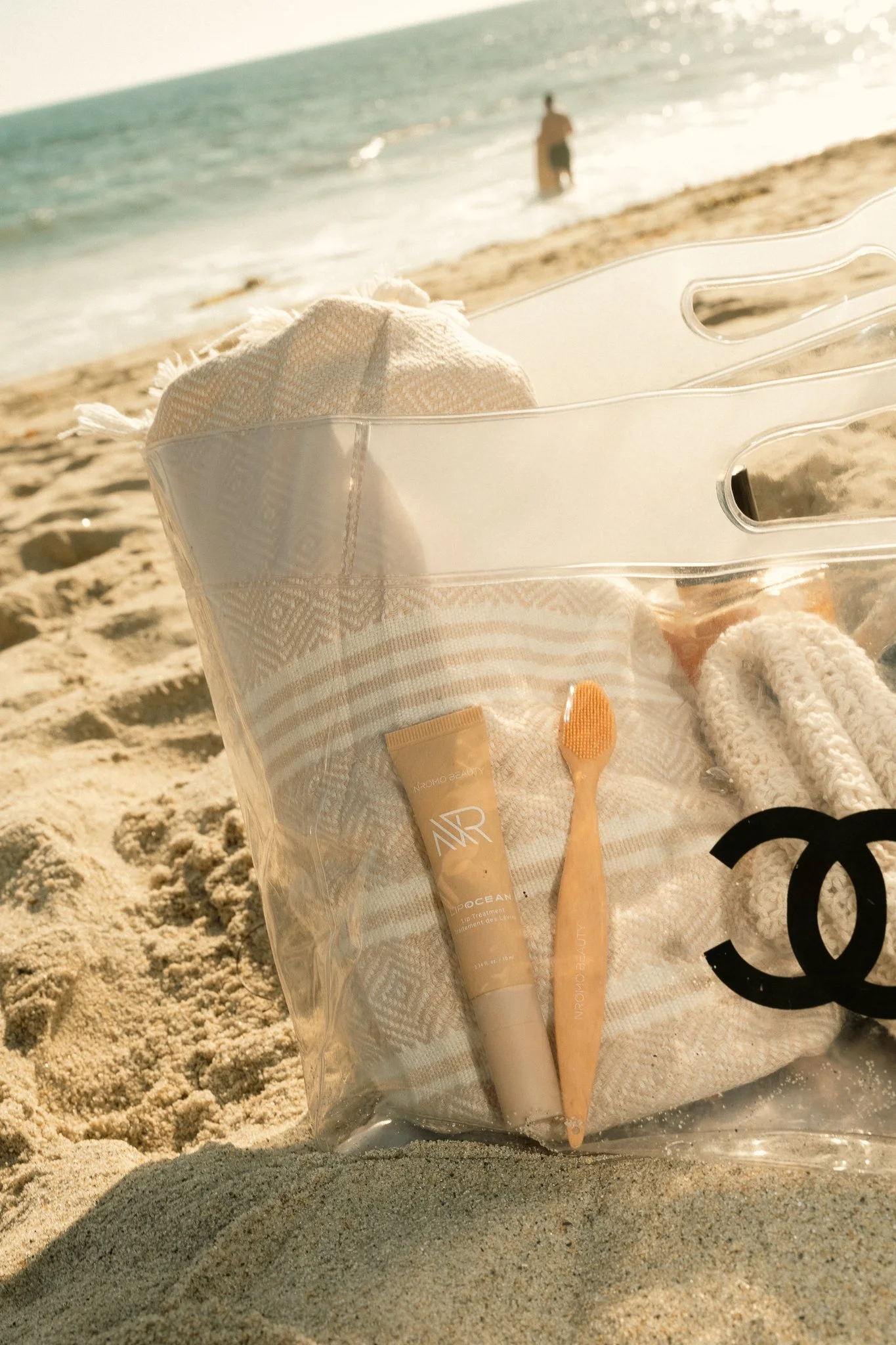 A clear plastic bag with beach essentials, including a tube of lip treatment, a wooden toothbrush, and a towel, sits on sandy beach with an ocean and a person wading in the water in the background.