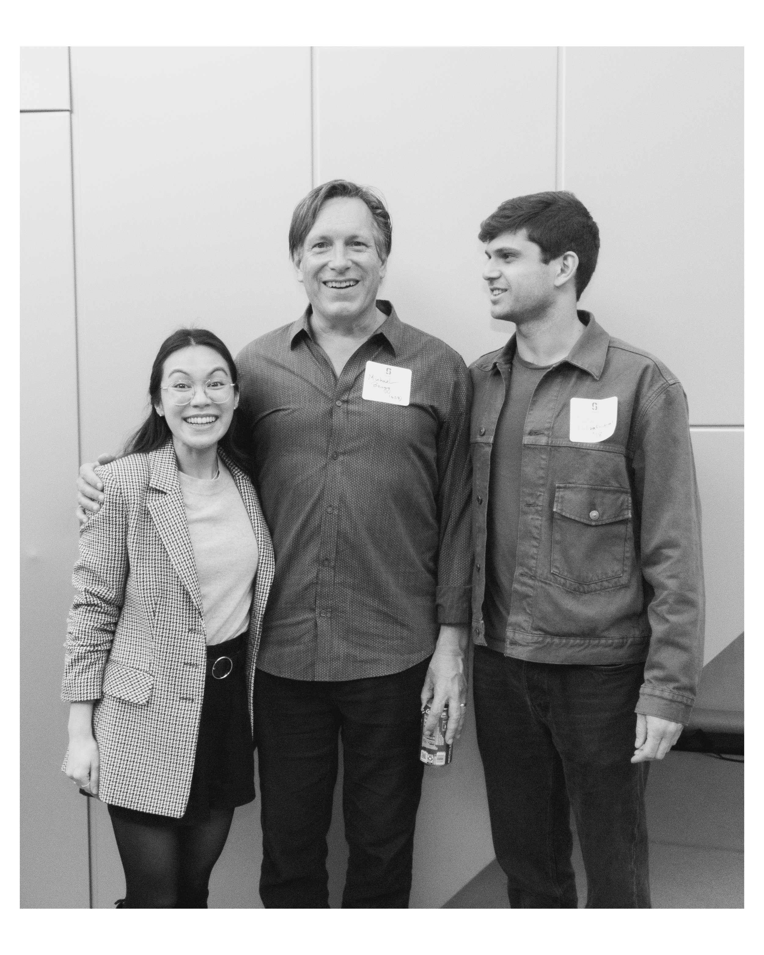 Three people, two men and one woman, smiling and standing close together in front of a plain wall. The woman on the left is wearing glasses and a checked blazer, the man in the middle has medium-length hair and is wearing a button-up shirt with a nam