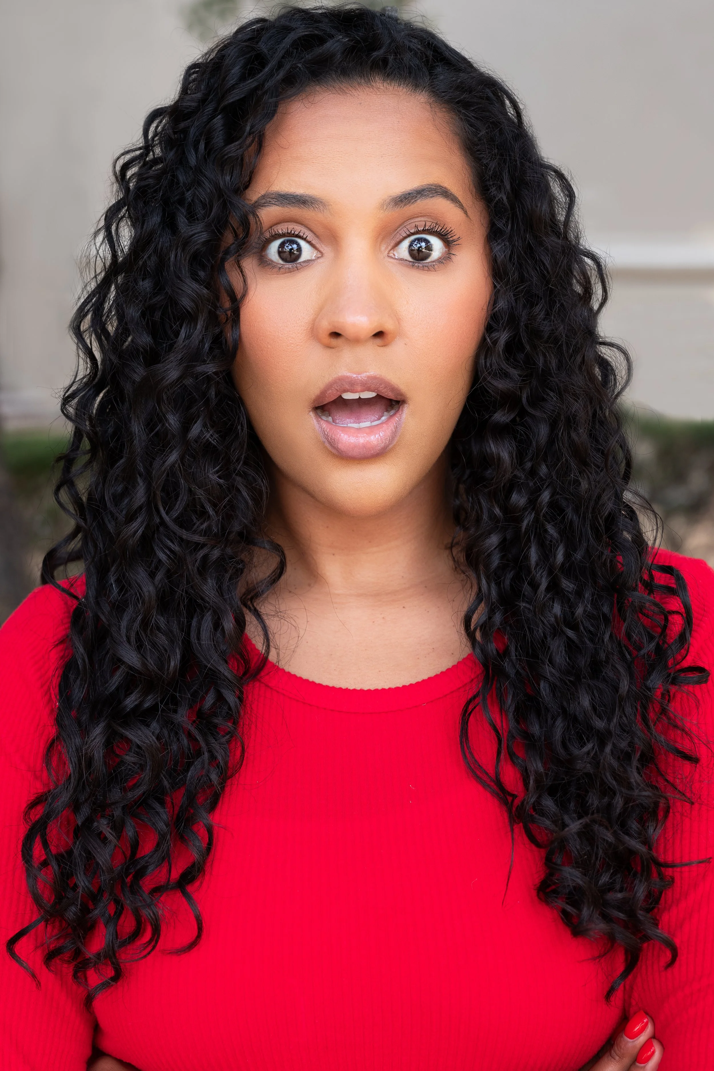 A woman with long black curly hair wearing a red top, her mouth open, expressing surprise or shock.