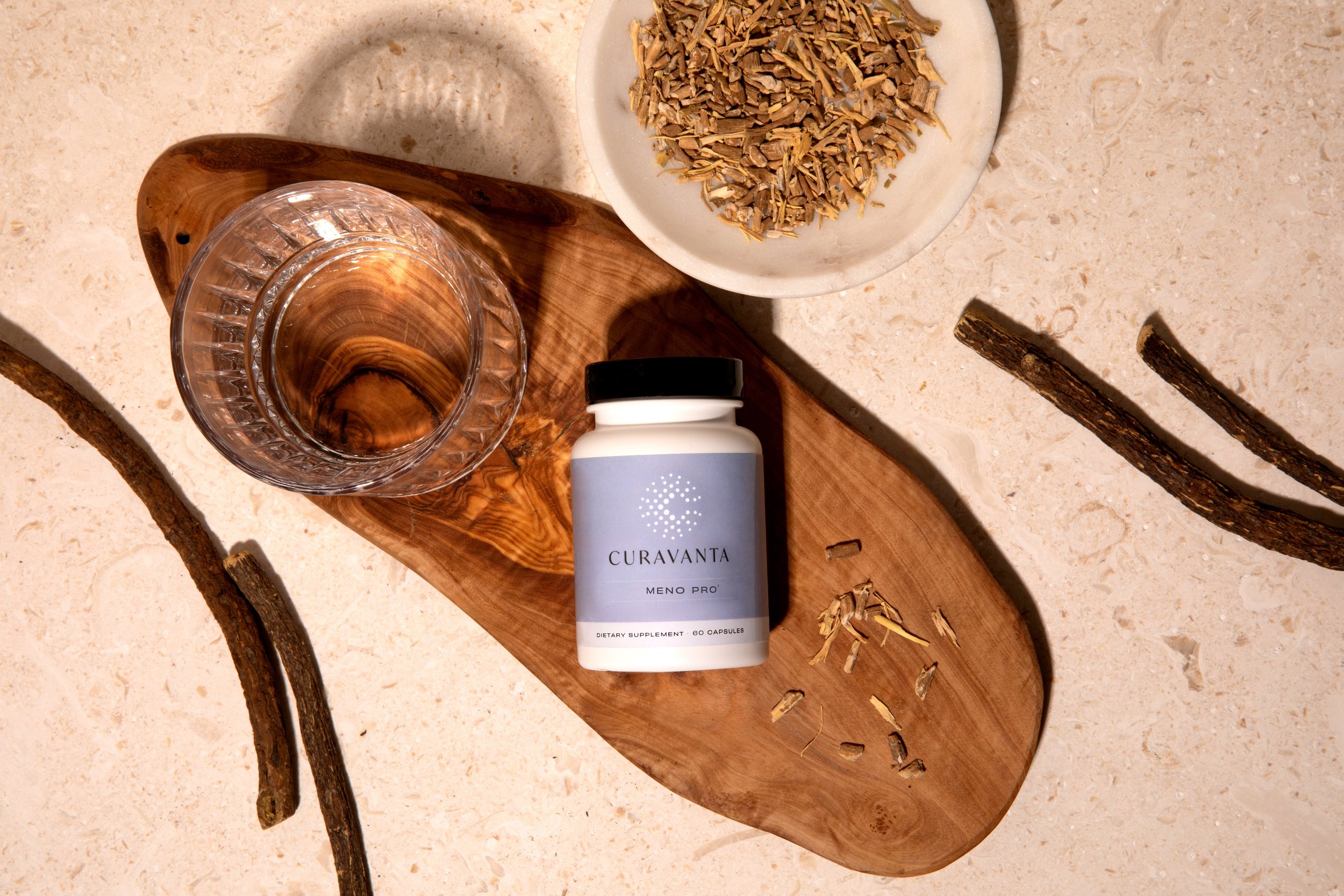 A glass of water, a bottle of Curavana Meno Pro dietary supplement, a wooden tray, a white bowl filled with dried herbs or plant material, several small sticks or twigs on the surface, and some loose plant material scattered around.
