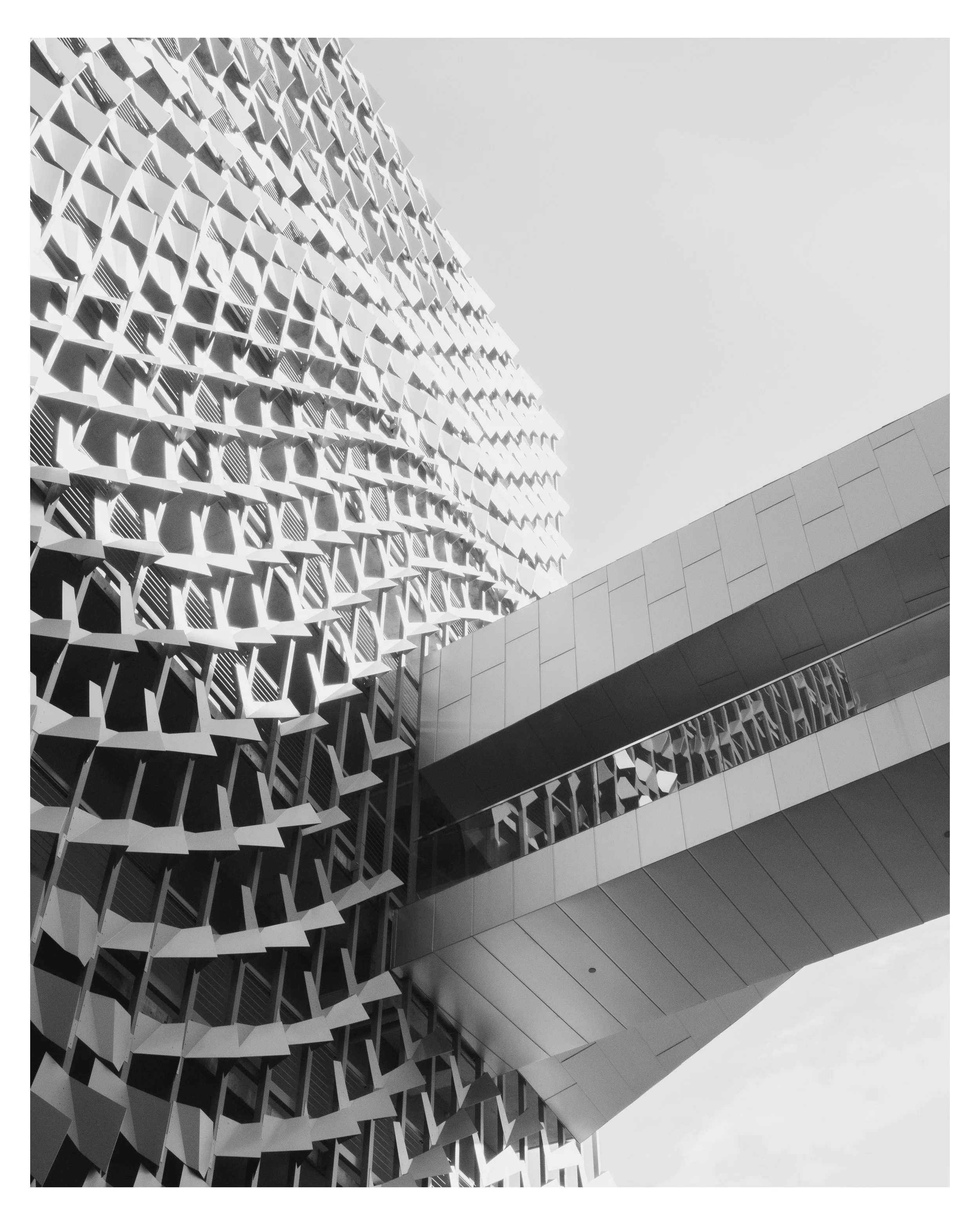 Black and white photo of a modern building with a distinctive geometric grid pattern on one facade, featuring horizontal and vertical elements creating a lattice design, and another section with smooth, flat surfaces, against an overcast sky.