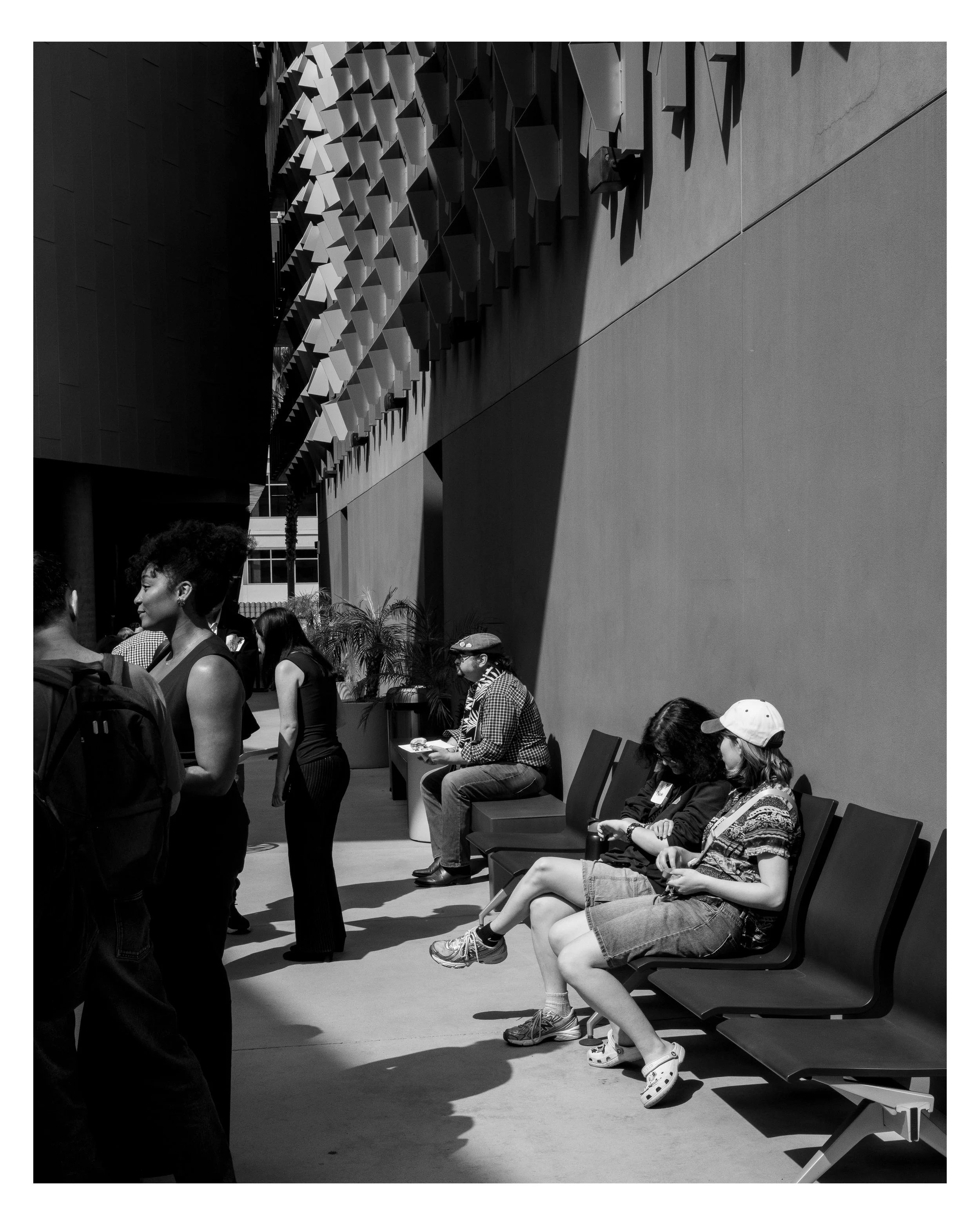 People sitting and standing outside a modern building with a unique facade pattern, some engaged in conversations, others waiting or looking at their phones.