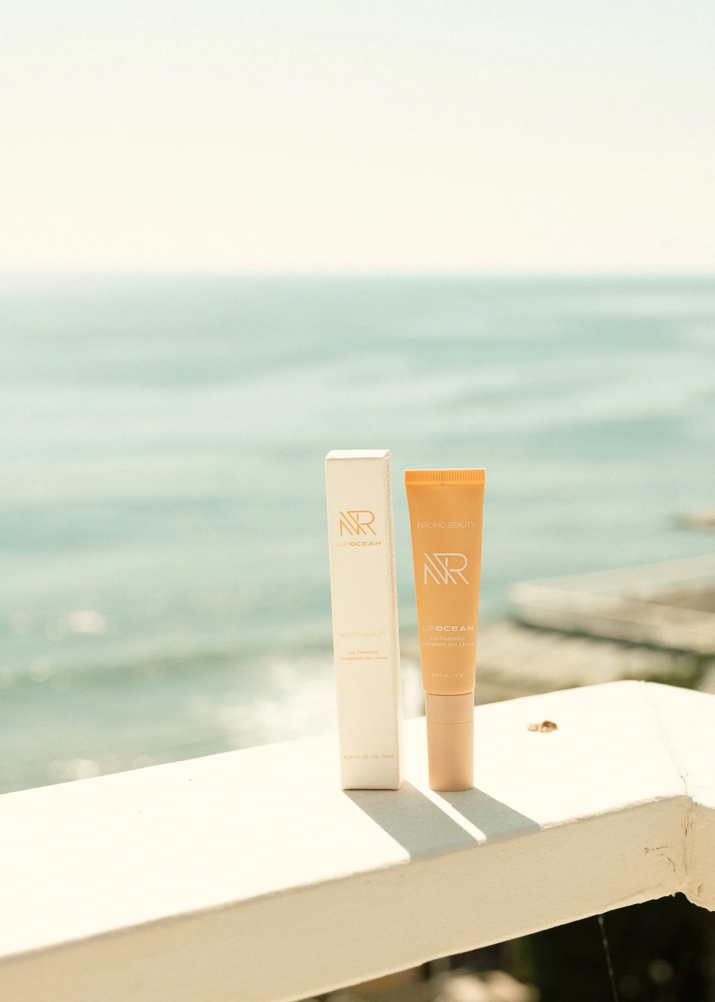 A tube of NAR Lip Ocean balm and its packaging on a windowsill with ocean and sky in the background.