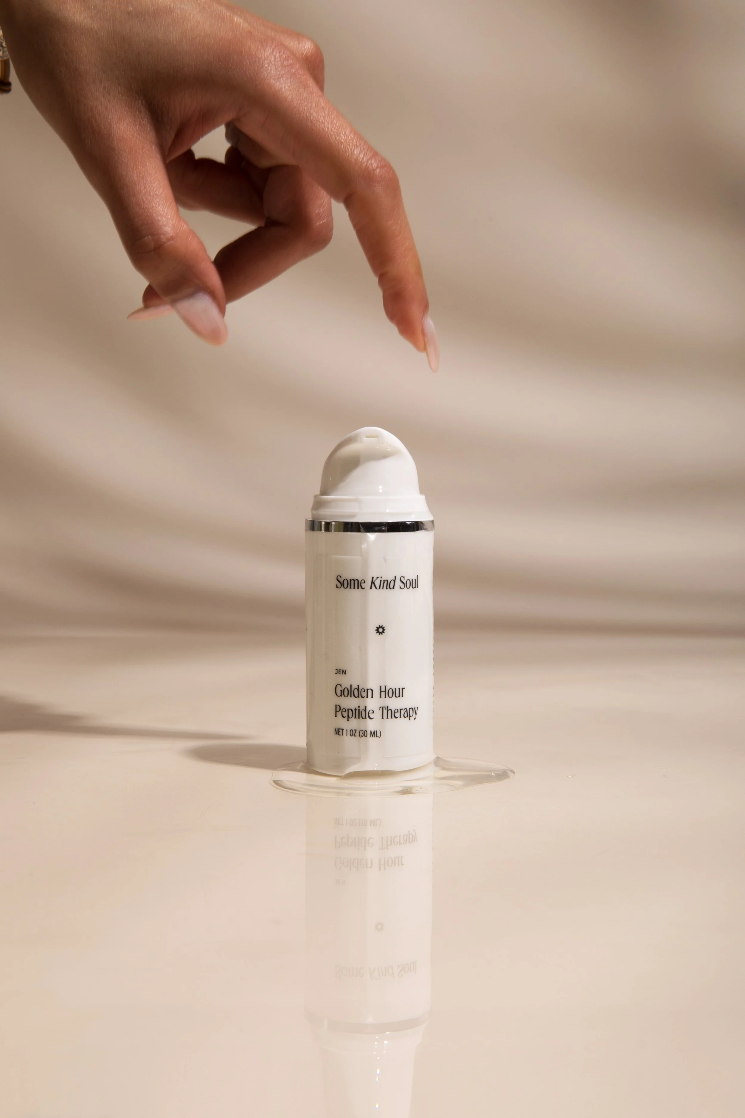 A hand with long, manicured nails reaches down to spray a white bottle labeled 'Some Kind Soul Golden Hour Peptide Therapy' onto a reflective surface.