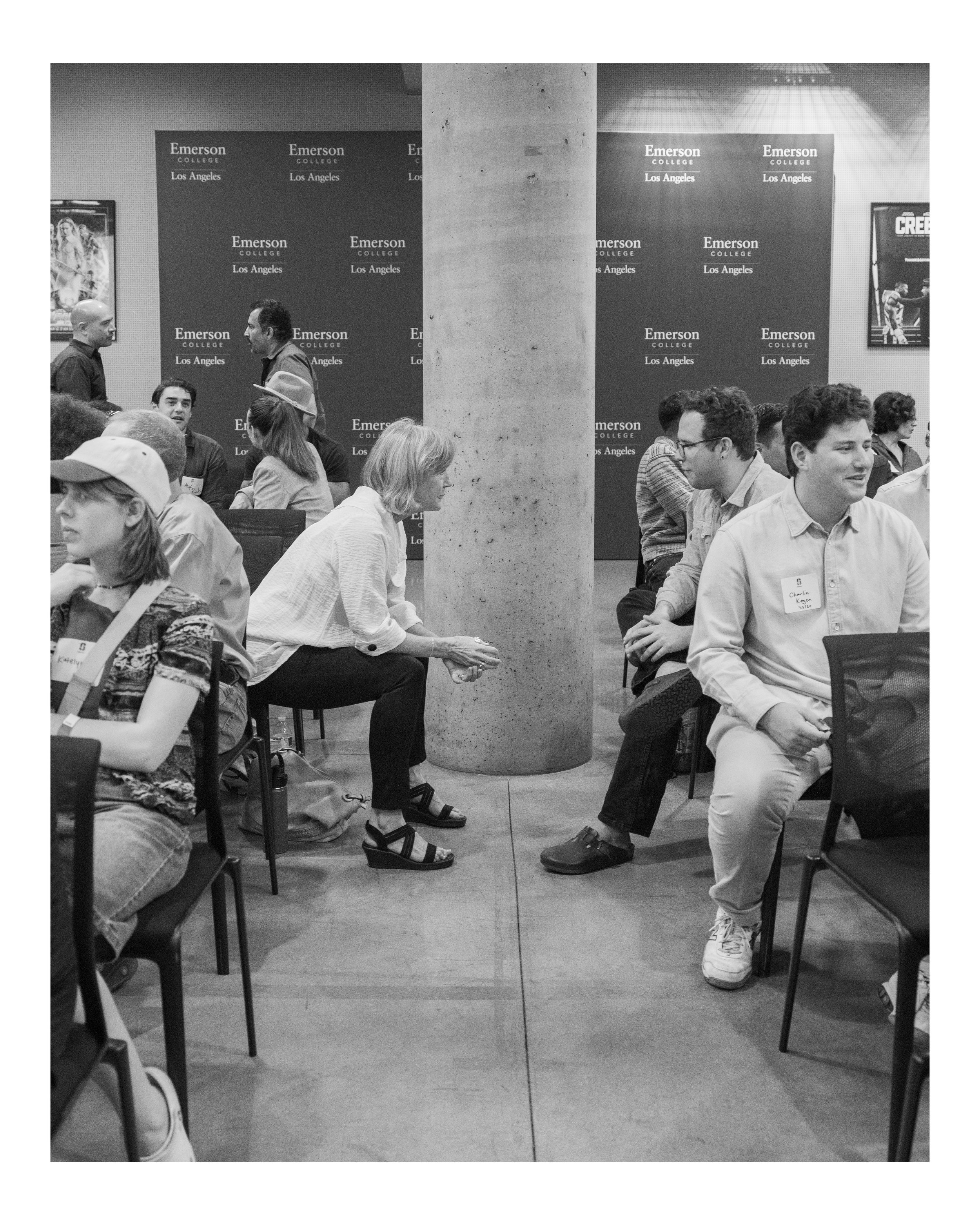 People sitting and talking at an event with a backdrop reading "Emerson College Los Angeles". A concrete pillar divides the scene, with individuals engaged in conversations, some wearing name tags.