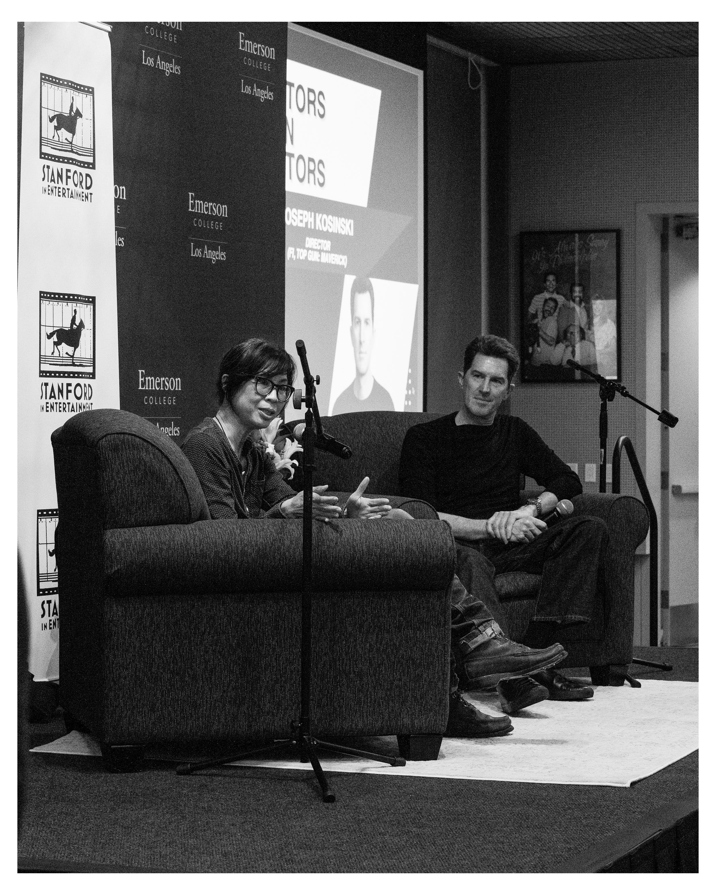 A panel discussion at Stanford in Entertainment, with three people seated on a stage. A woman in glasses is speaking into a microphone, a man on her right is holding a microphone, and another person’s head is visible in the foreground. There are bann