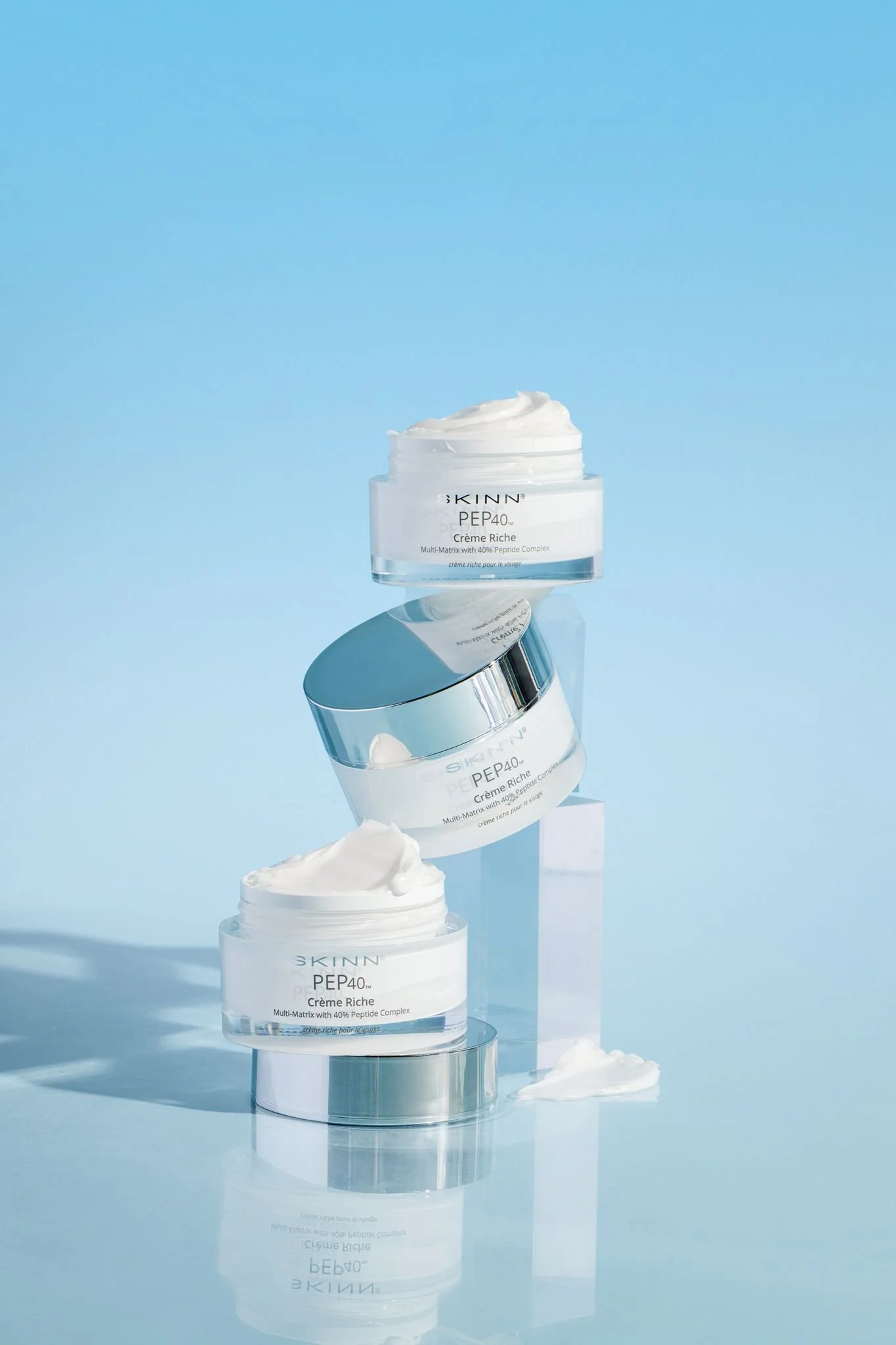 A display of skincare products, specifically jars of SKINN PEP40 Creme Riche, arranged on mirrored stands against a light blue background with reflections visible.