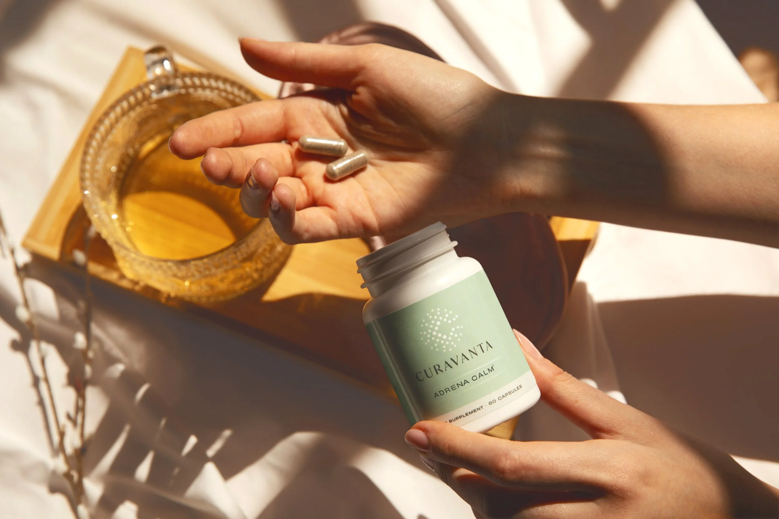A person's hand holding a bottle of Curavanta Adrena Calm supplement capsules, with a few capsules in the palm, near a decorative glass candle holder on a wooden surface, with sunlight casting shadows.