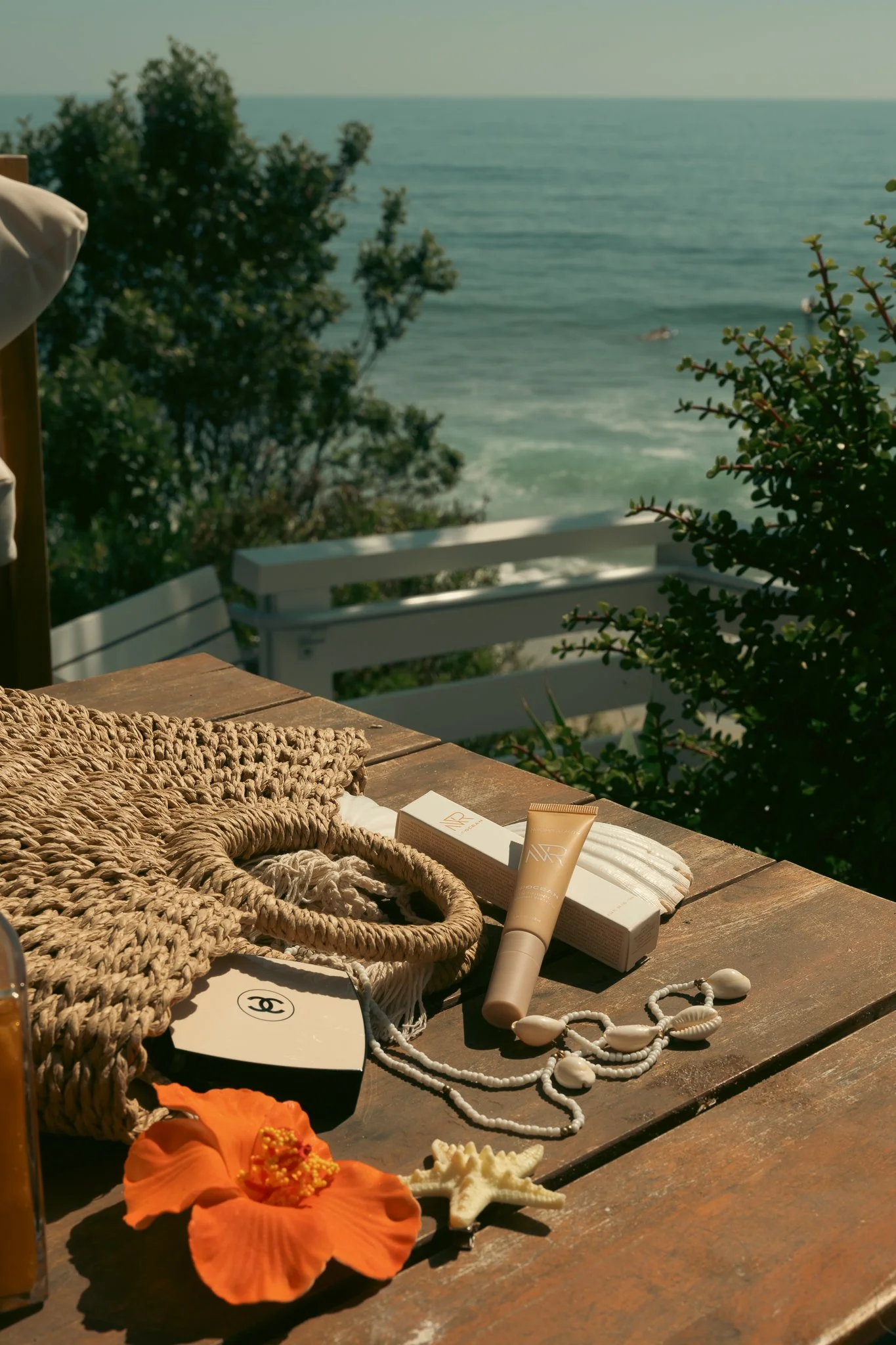 A wooden table holding various items including a loofah, Chanel cosmetics, a seashell necklace, orange hibiscus flower, starfish, hair ties, and a tube of skincare product, with a view of trees, the ocean, and a distant boat in the background.