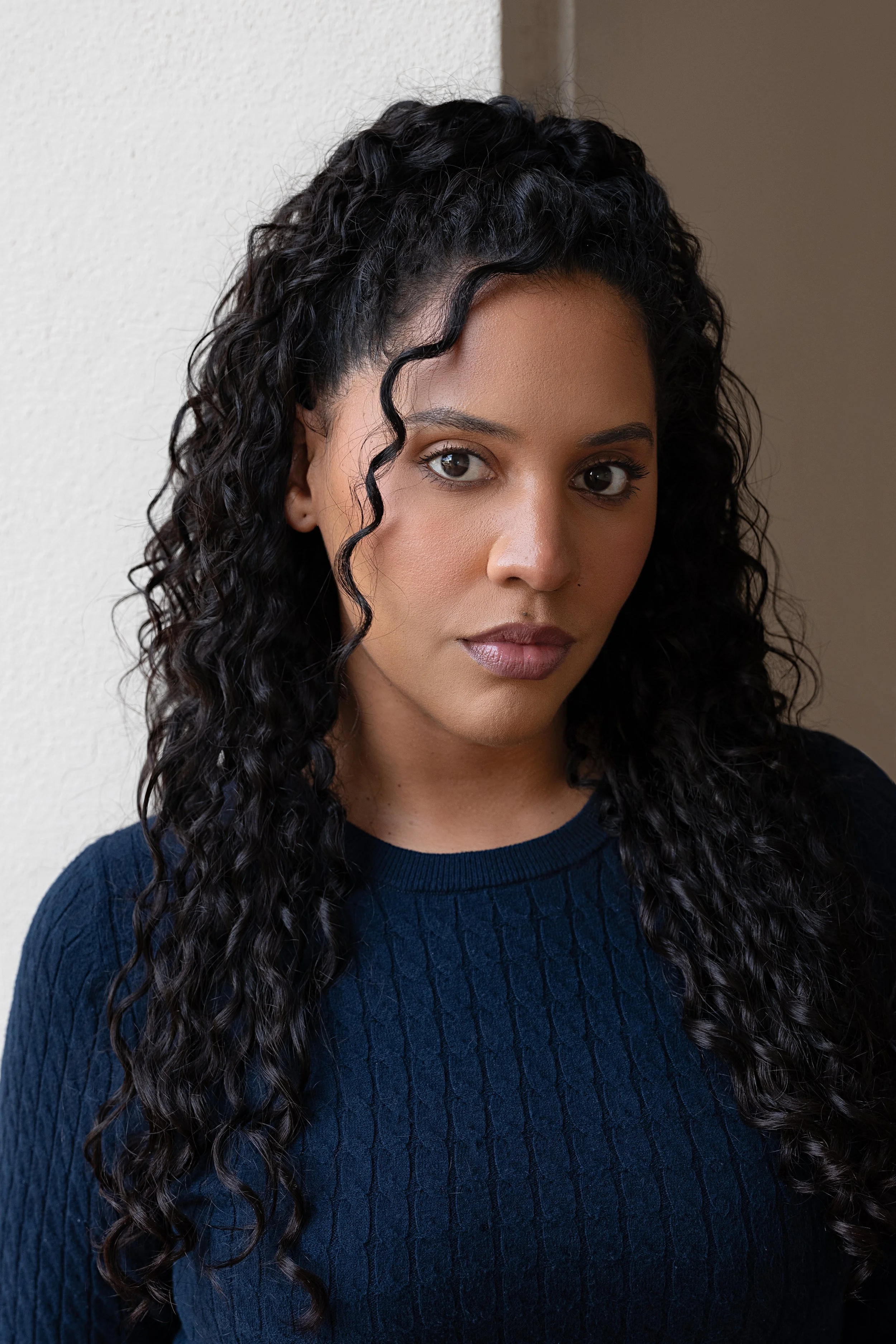 Close-up of a young woman with long, curly dark hair, wearing a navy blue sweater, standing against a neutral background.