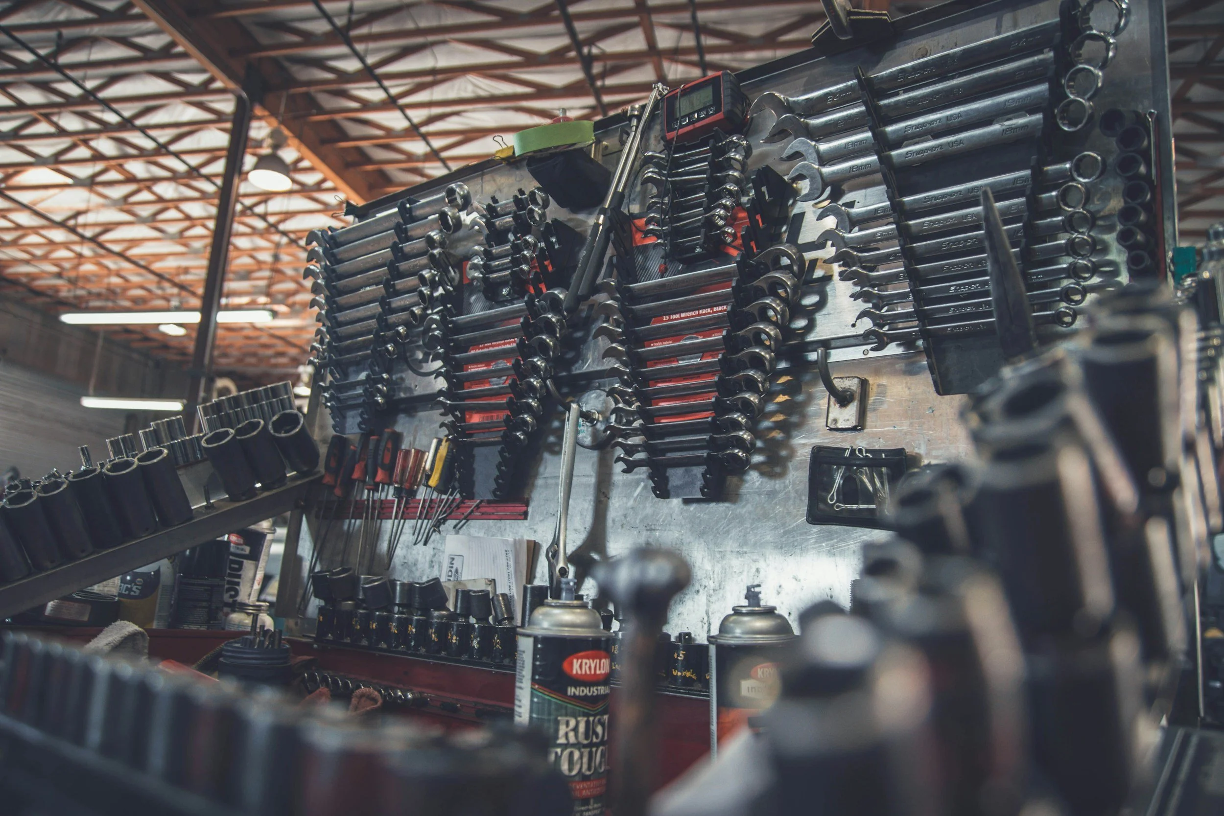 A workbench in a workshop holding various tools such as wrenches, screwdrivers, and bottles of lubricant, with a warehouse ceiling overhead.