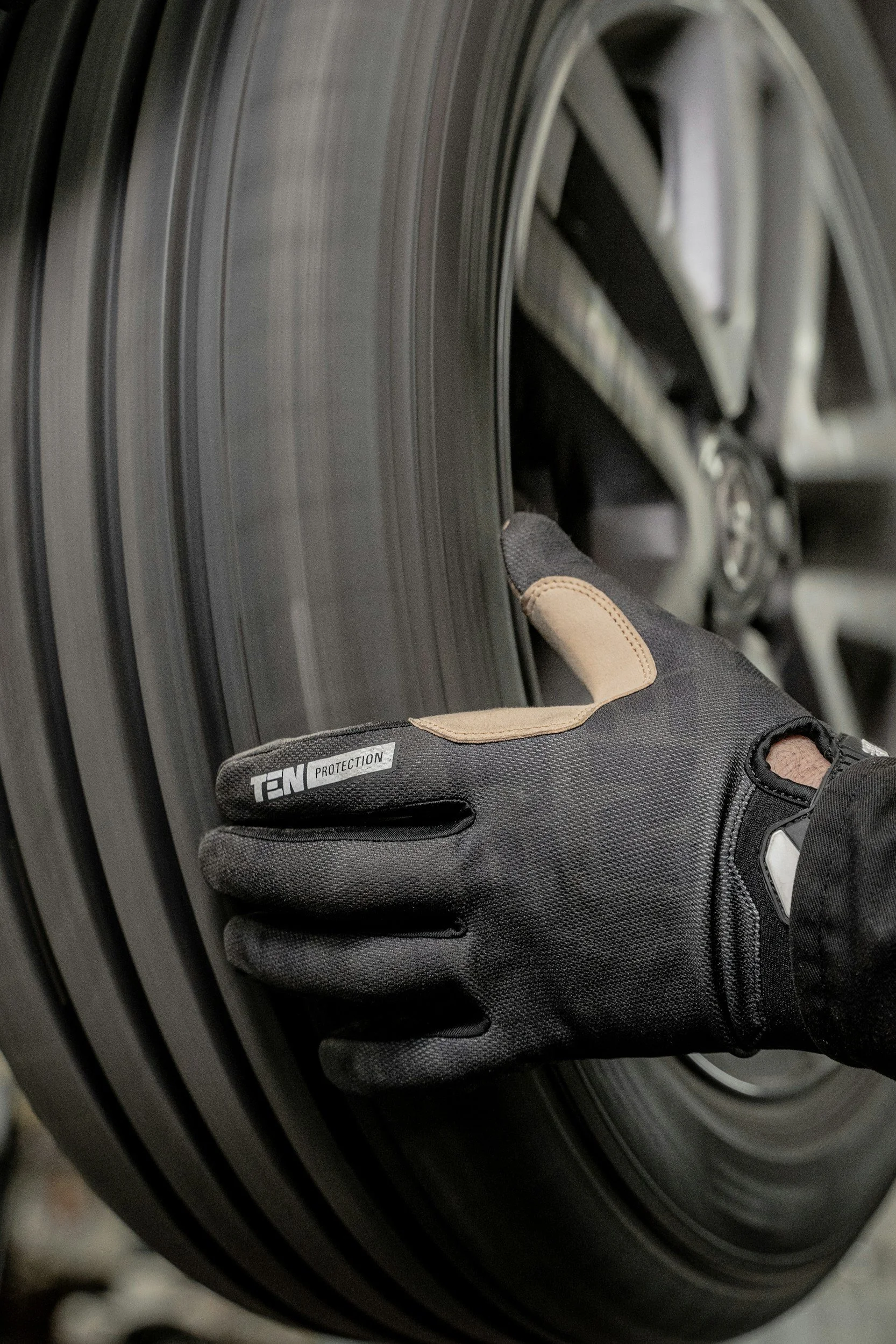 Close-up of a person's hand wearing a black glove with tan accents, touching the rim of a vehicle tire.