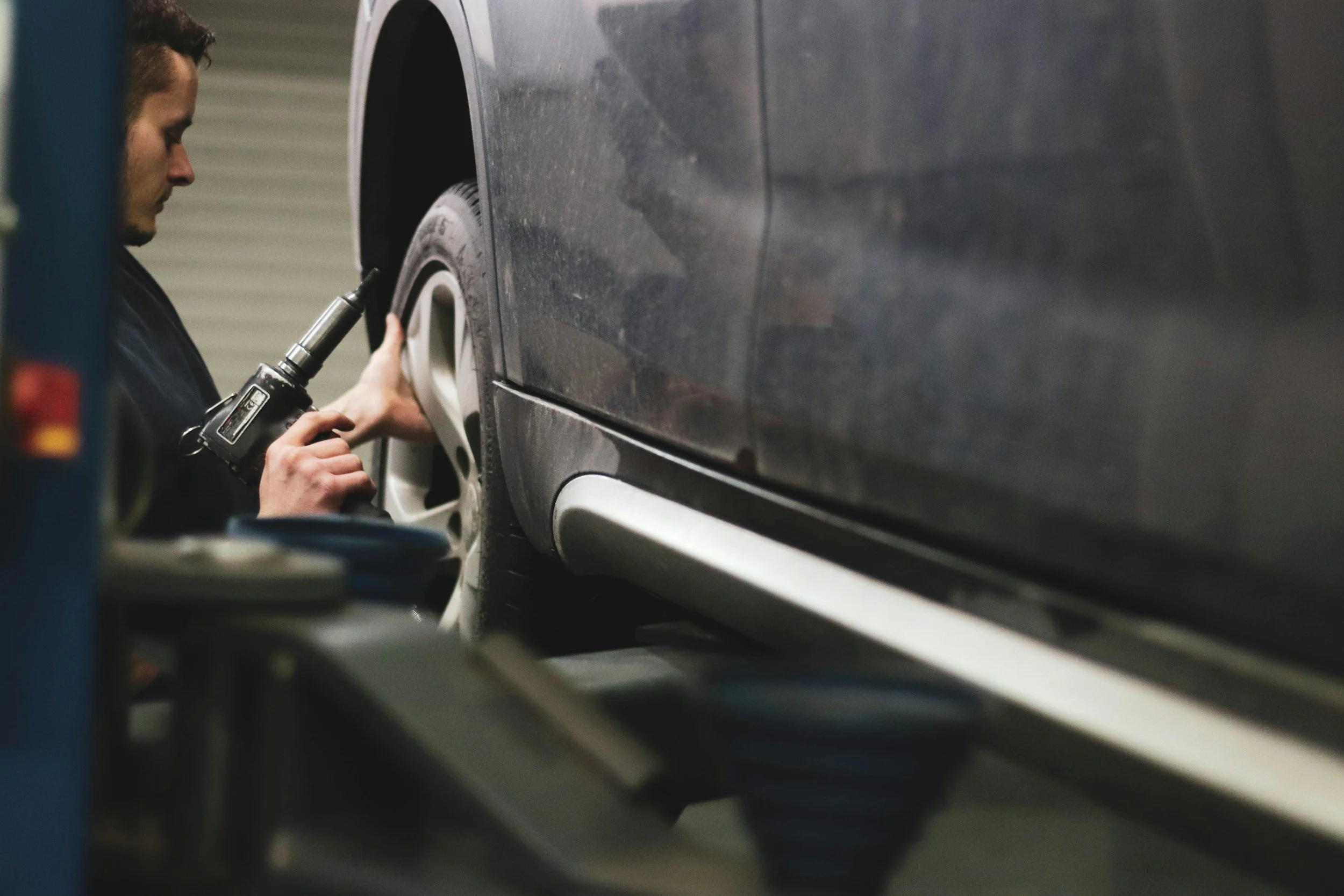 A mechanic using a cordless drill to work on the rear wheel of a dark-colored car in an indoor garage.