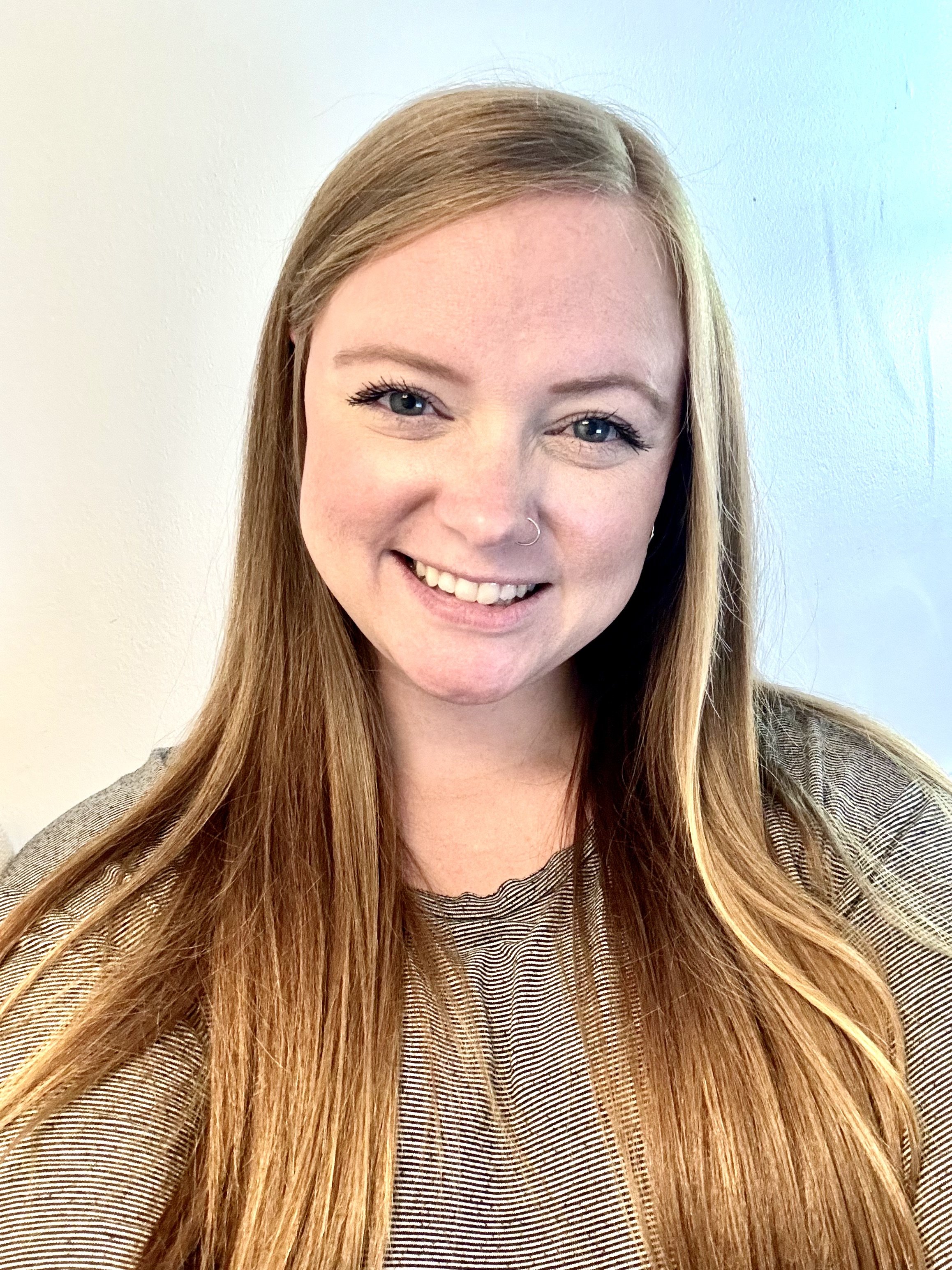 A woman with long, straight red hair, a nose ring, and blue eyes smiling at the camera, wearing a striped shirt, in front of a plain wall.
