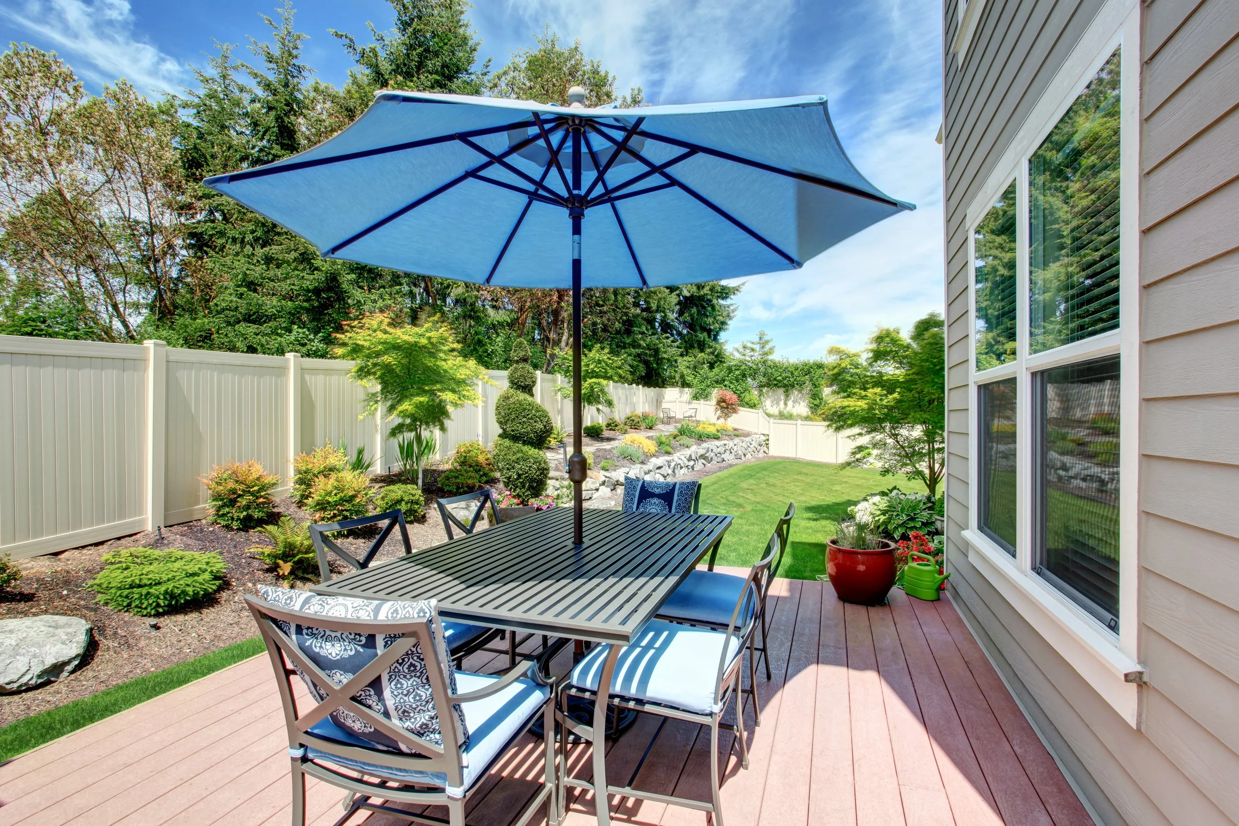 A backyard patio with a black metal table and six chairs, shaded by a large blue umbrella. The yard has a well-maintained lawn, decorative bushes, and a white privacy fence, with trees and landscaping in the background.