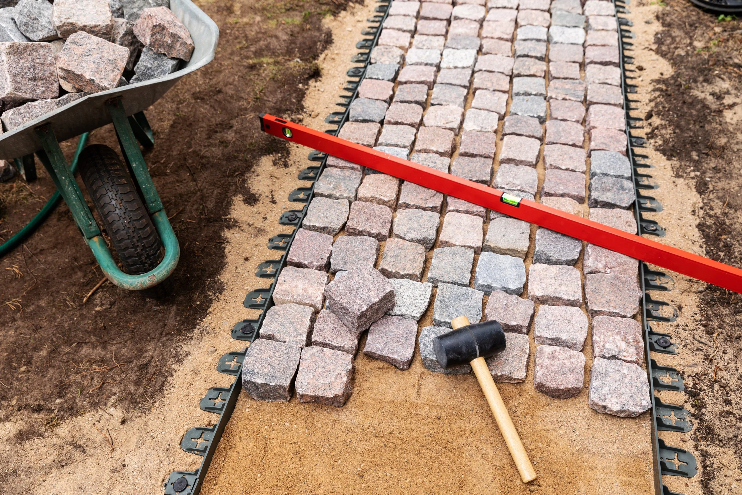 A cobblestone pathway under construction with a level tool placed on top, a hammer resting on the ground, and a wheelbarrow containing stones nearby.