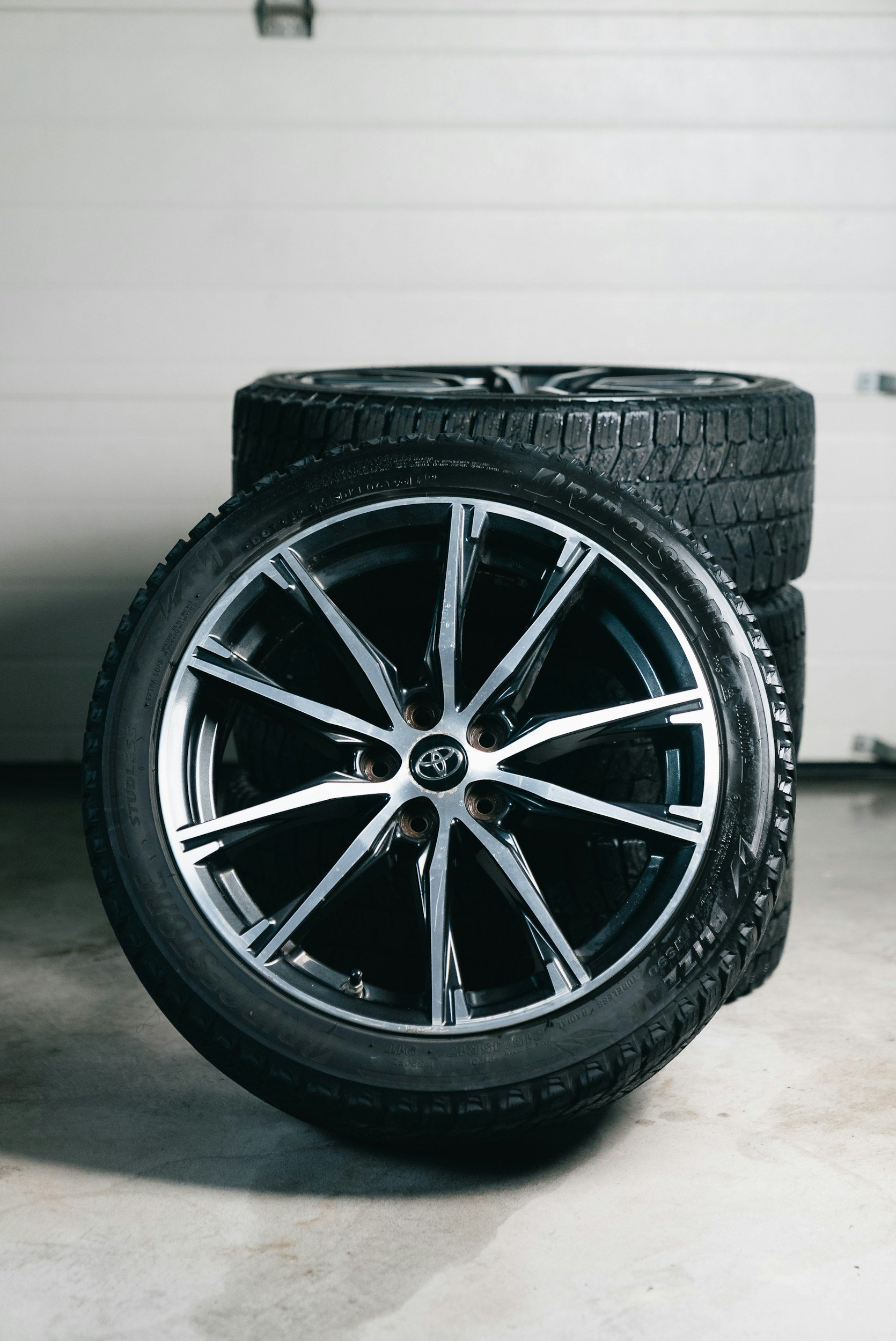 A close-up of a black and silver Toyota alloy wheel with a tire mounted, sitting on a concrete floor with two additional tires stacked behind it and a white garage door in the background.