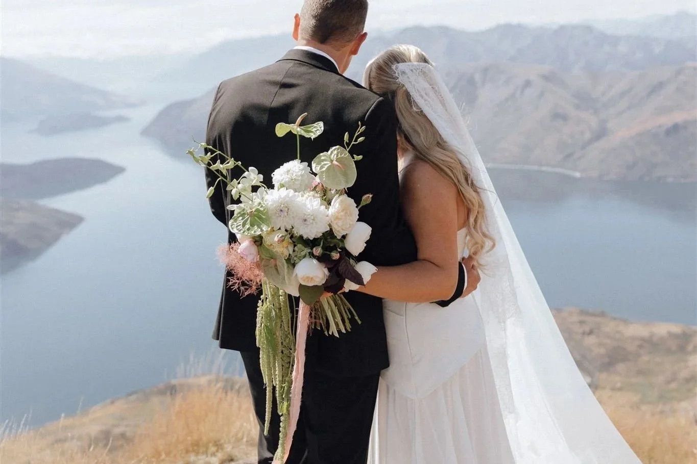 A bride and groom standing together with a scenic lake and mountains in the background, the bride holding a large bouquet of white and pink flowers.