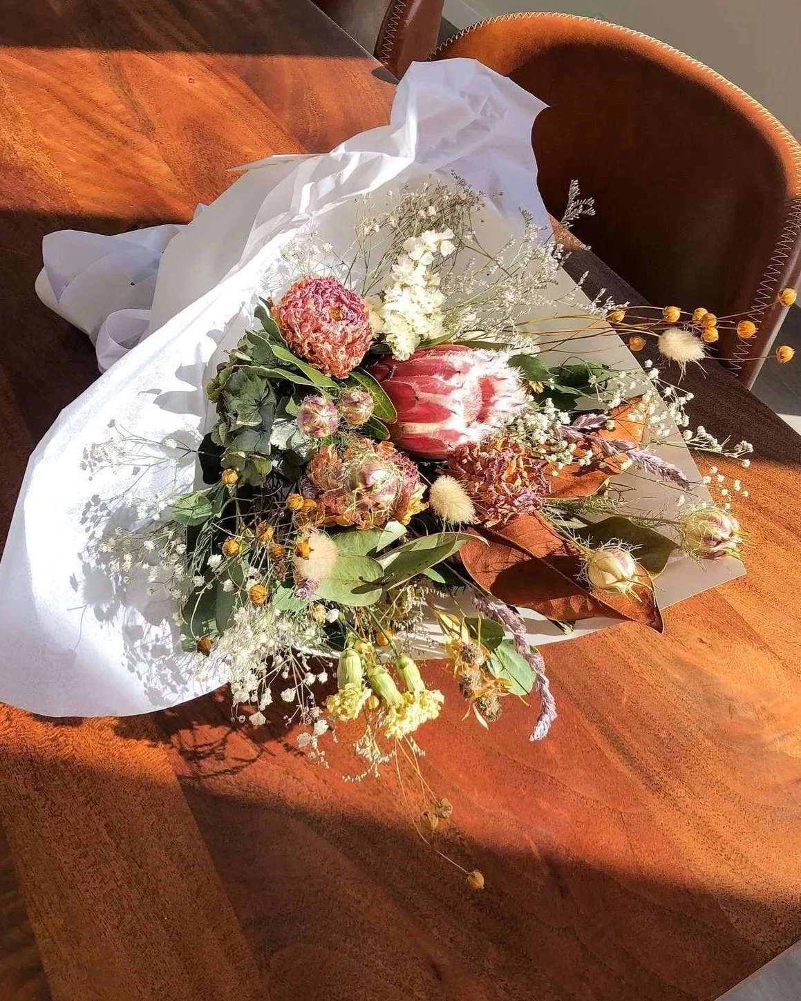 A bouquet of mixed flowers on a wooden table, wrapped in white and brown paper, with a variety of flowers including protea, baby's breath, and other wildflowers.
